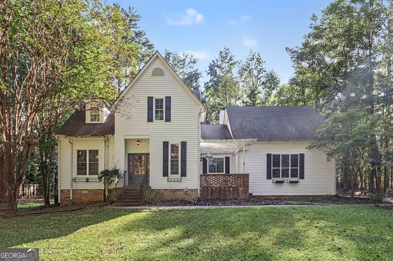 Cream-sided house with dark shutters and brick accents, surrounded by mature trees and a broad lawn.