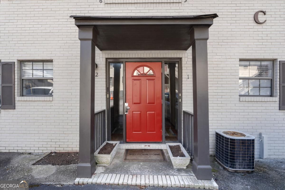 White brick building entrance with a bright red door beneath a dark covered porch.