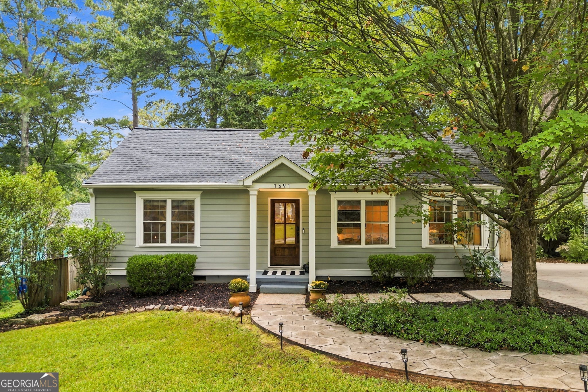 Gray-green single-story house with white trim, shaded by mature trees and approached by a curved stone walkway.
