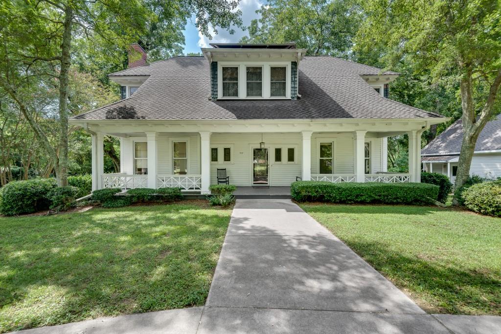 White two-story house with a broad covered porch and central dormer, framed by mature trees.