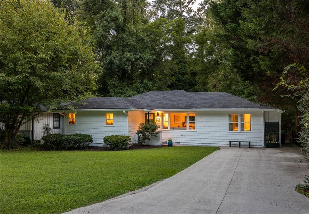 White single-story house with warmly lit windows, a broad lawn, and a concrete driveway beneath mature trees.
