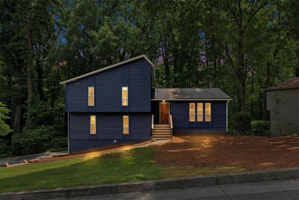Blue split-level house with a warmly lit entry, sloped lawn, and wooded surroundings.