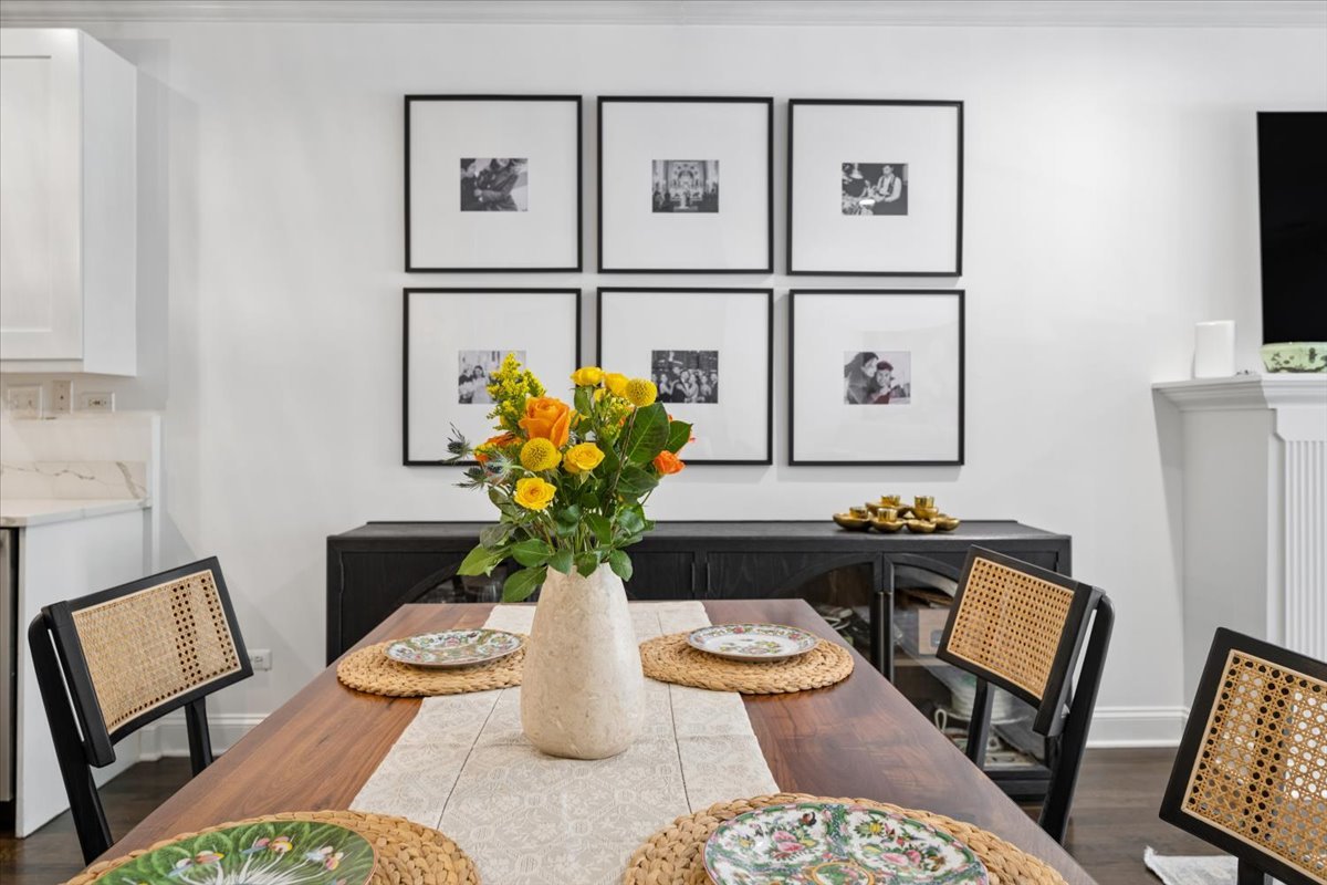 Dining area with a flower centerpiece, cane-back chairs, and framed photographs above a dark sideboard.