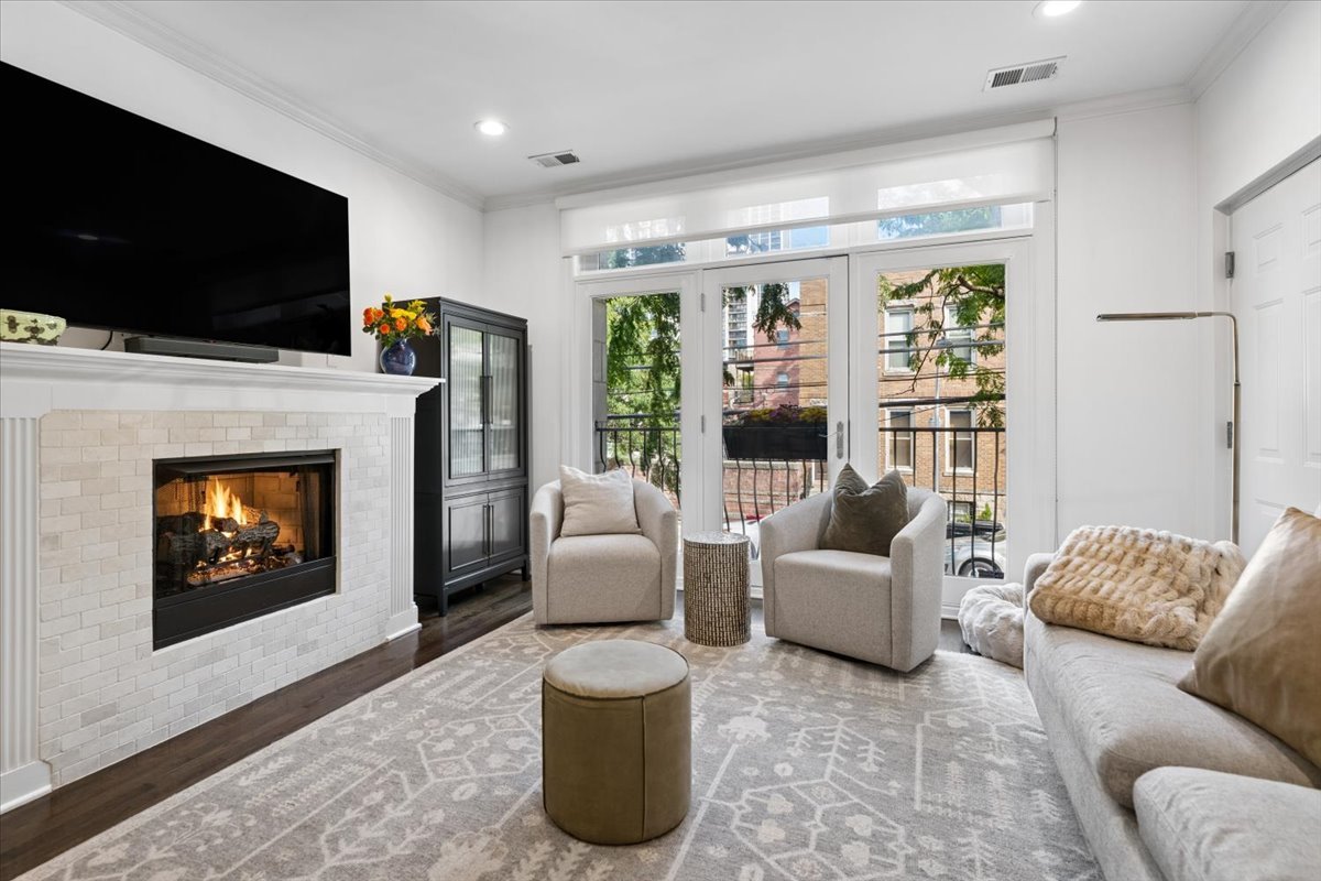 Living room with a white tile fireplace, pale seating, and balcony doors overlooking brick buildings.