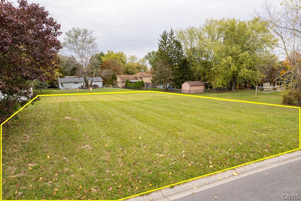 Grassy lot outlined in yellow beside a street, with mature trees and neighboring homes in the background.