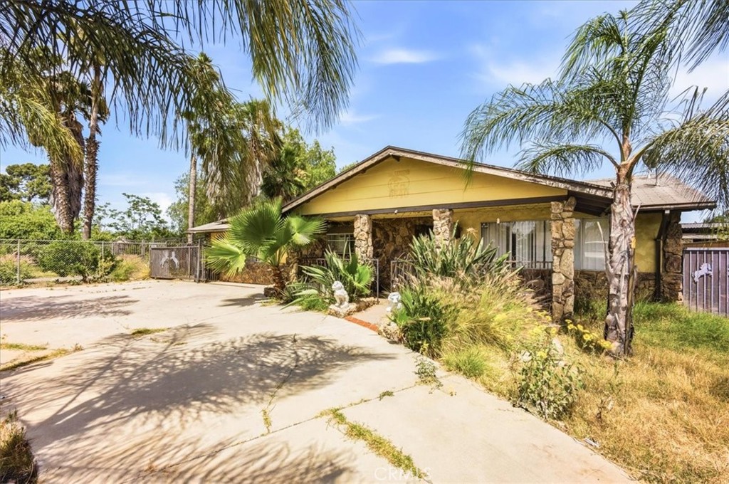 Single-story yellow house with stone columns, palm trees, and a broad concrete driveway.