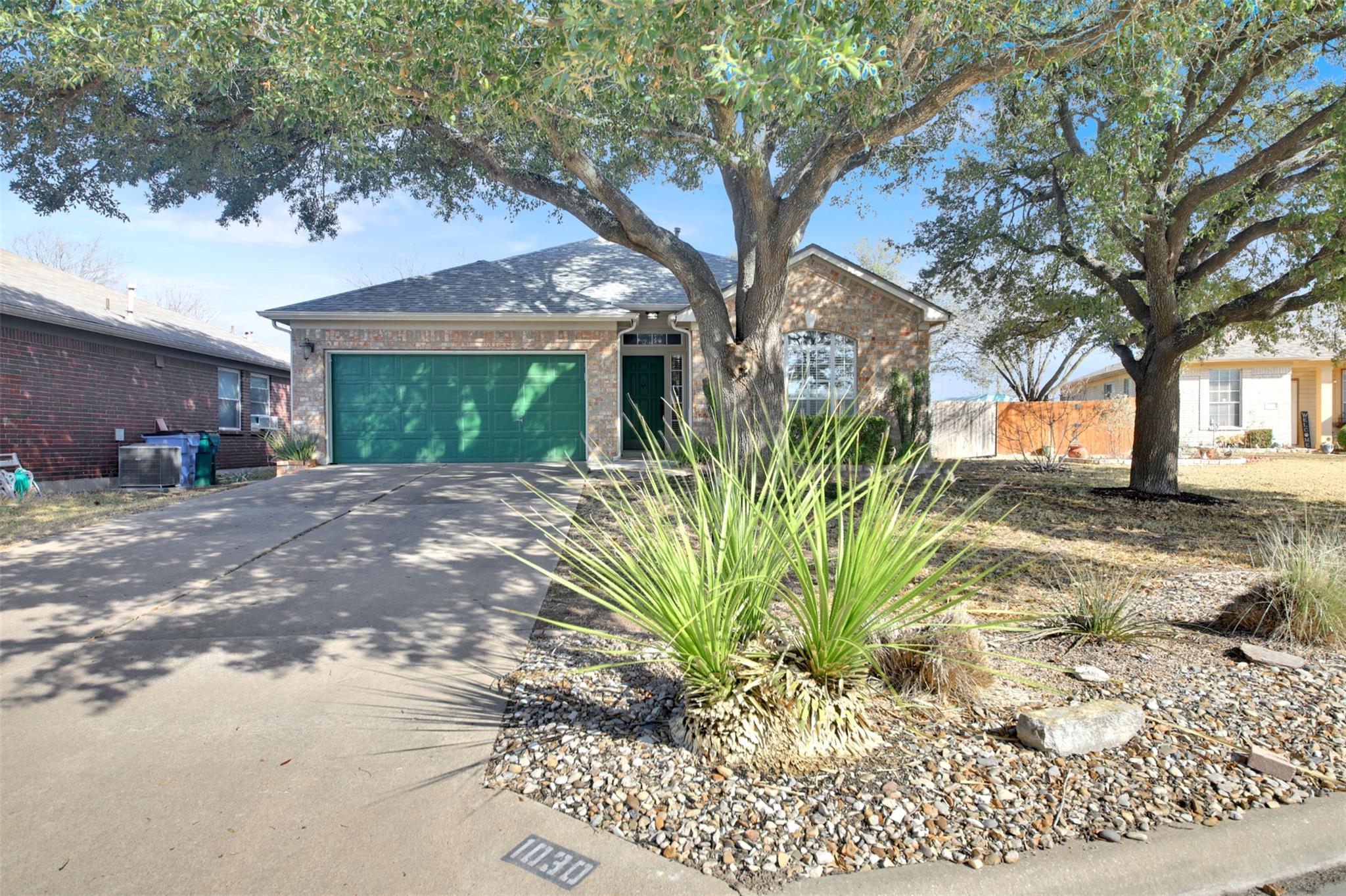 Brick house with green garage door and rock landscaping beneath large trees.
