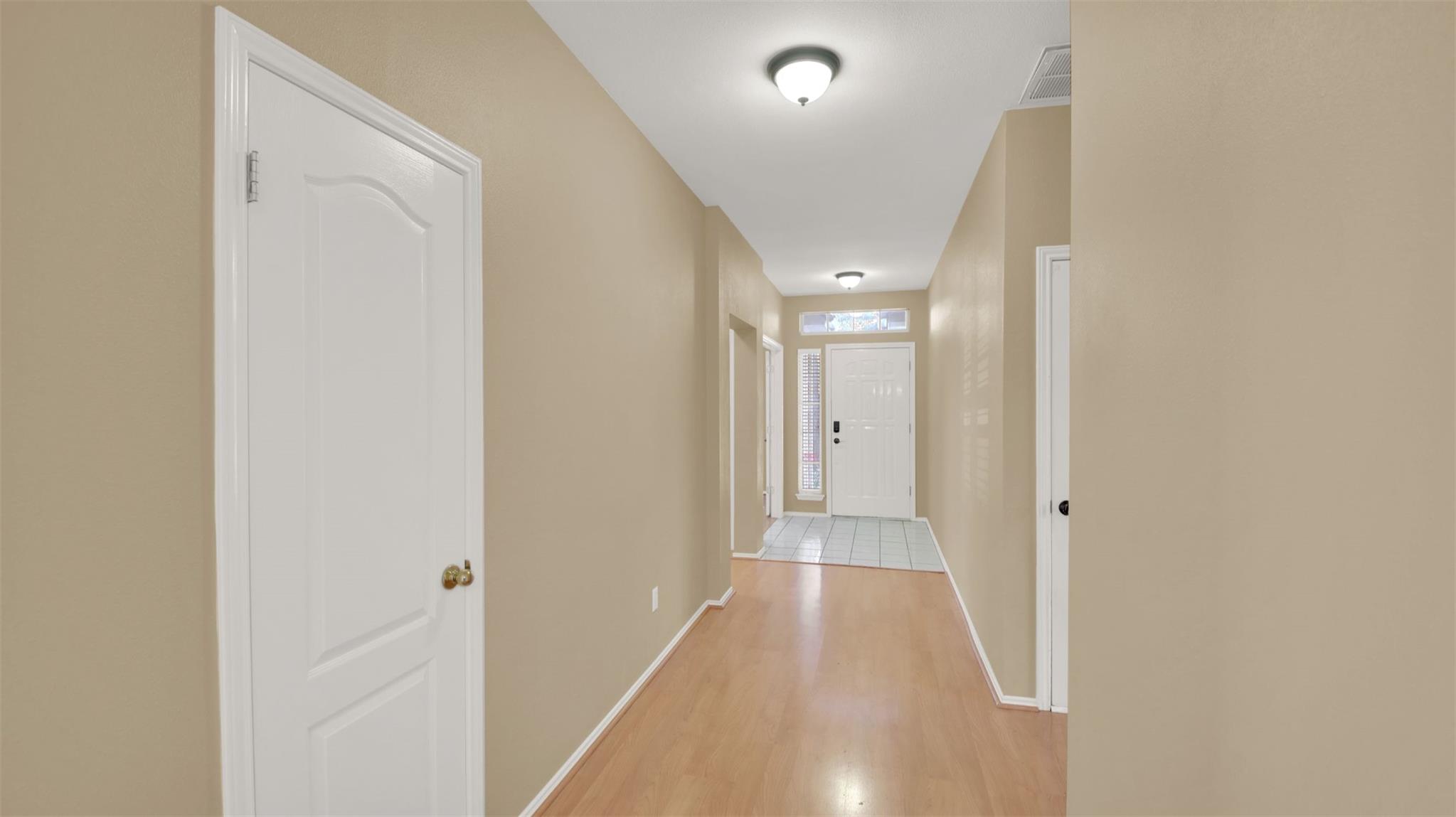 Entry hallway with light wood flooring leading to a tiled foyer and white front door.