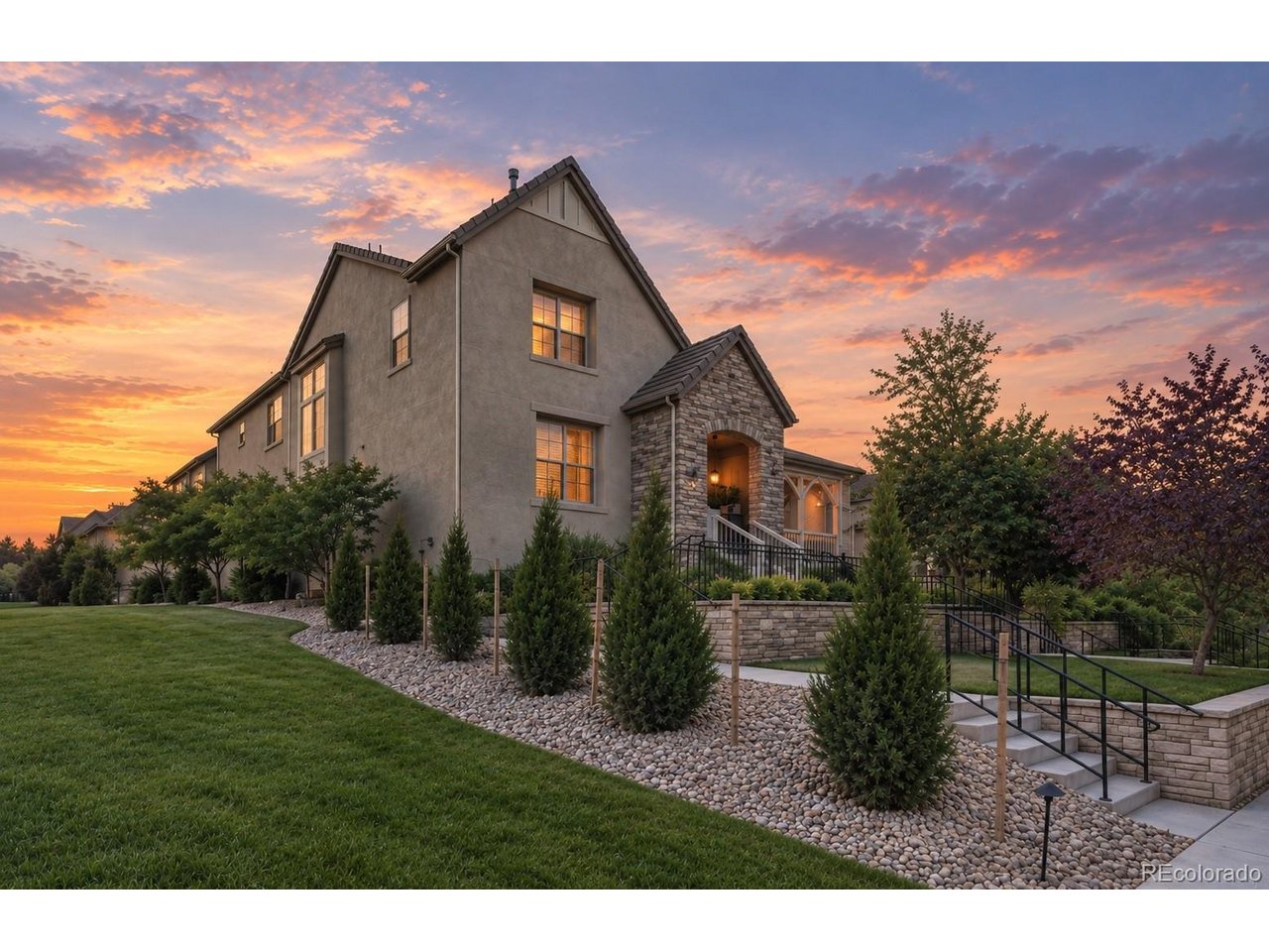 Two-story stucco and stone house with landscaped grounds beneath a pink-orange dusk sky.