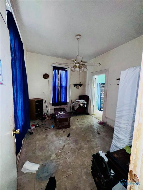 Cluttered living room with blue curtains, a ceiling fan, and scattered furniture and belongings.