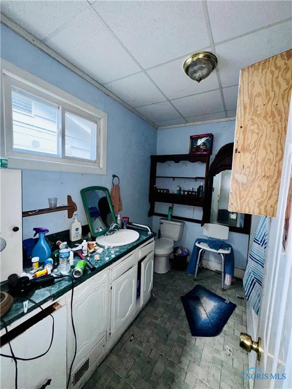 Bathroom with a long vanity, cluttered countertop, toilet, and small high windows.