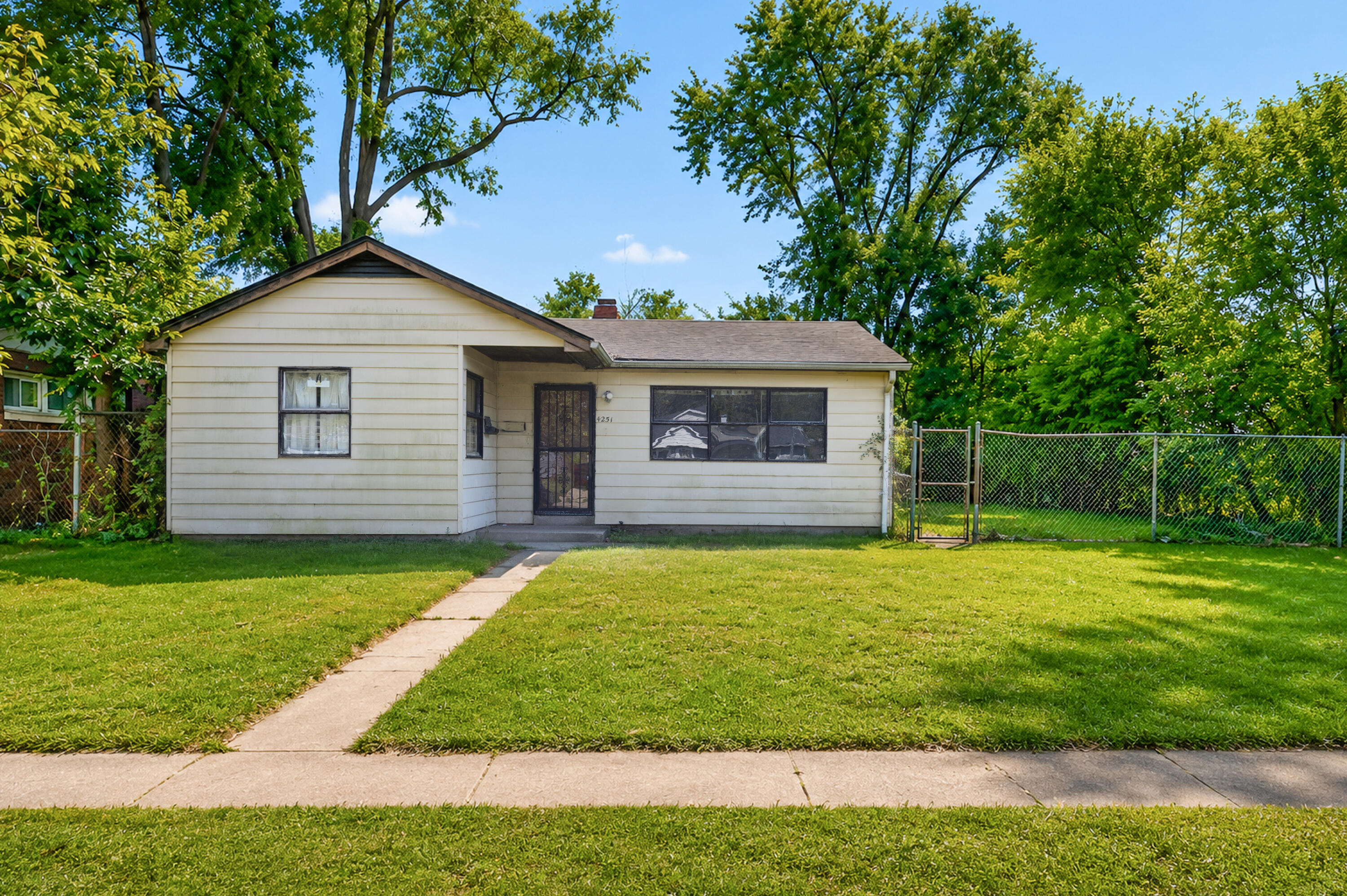 Single-story cream-sided house with a front lawn, walkway, chain-link fence, and mature trees.