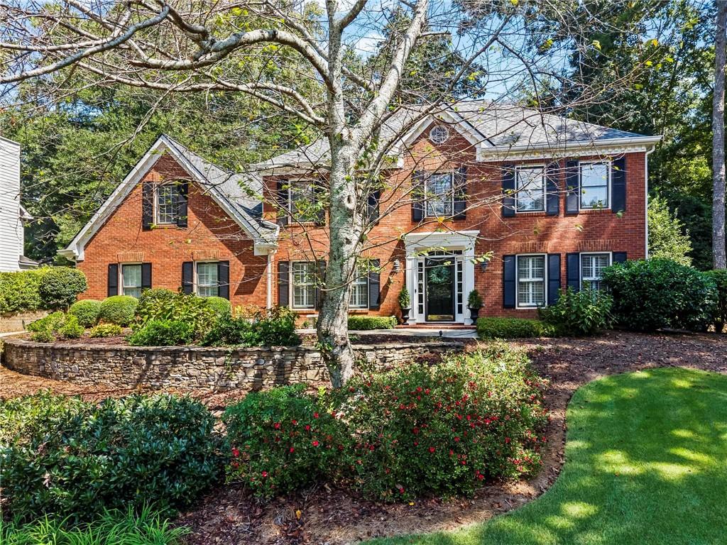Two-story red brick home with dark shutters, mature trees, and a landscaped stone retaining wall.