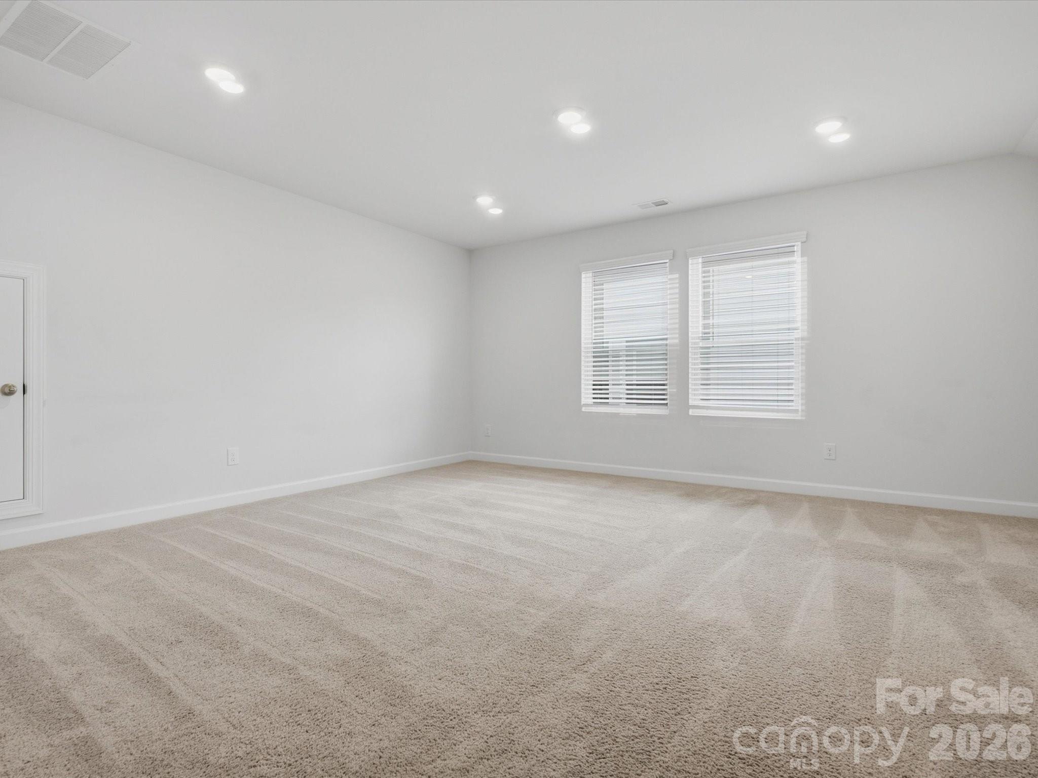 Empty carpeted room with white walls, recessed lights, and two windows with blinds.