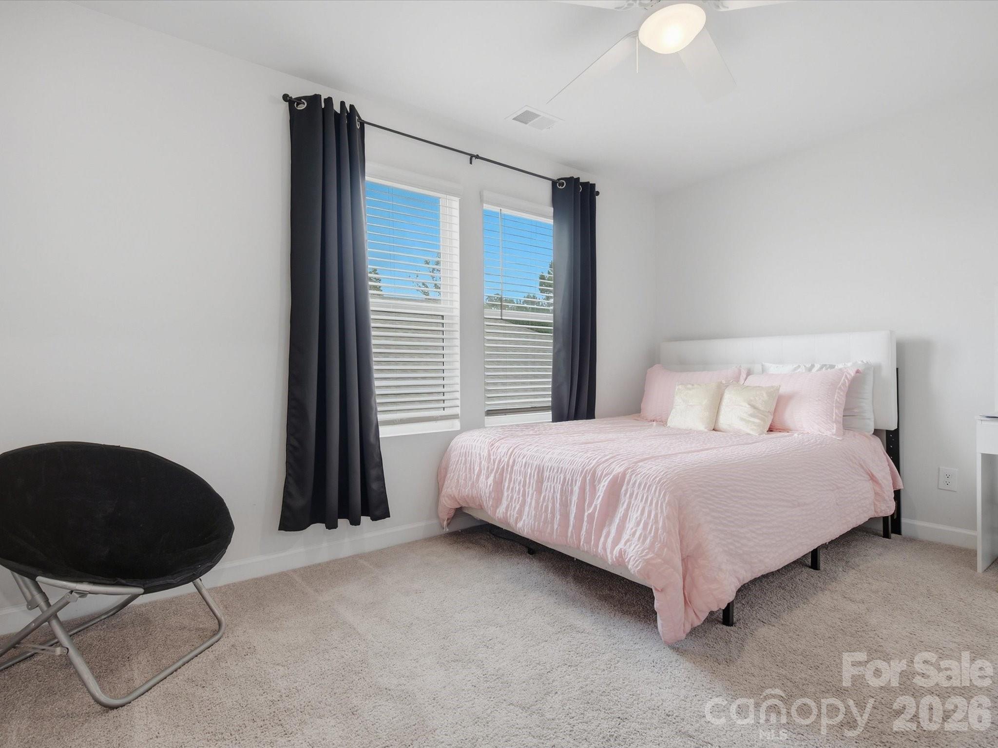 Staged bedroom with a pink bed, dark curtains, two windows, and a black lounge chair.
