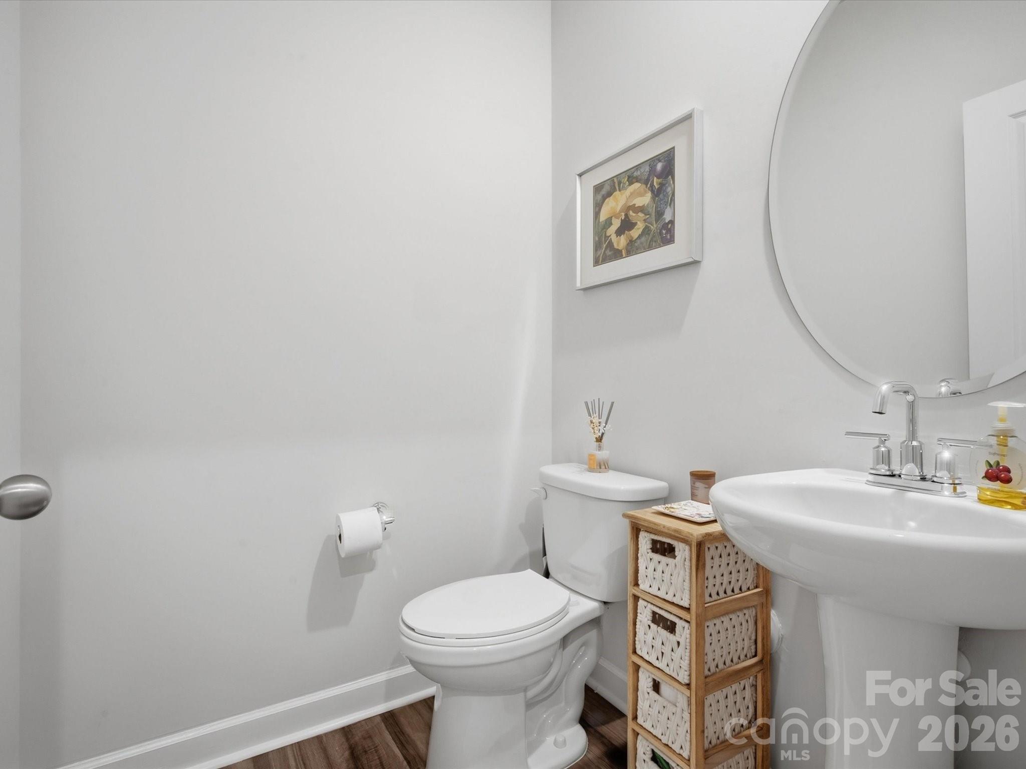 Bathroom with a white toilet, pedestal sink, large round mirror, and wooden basket storage.