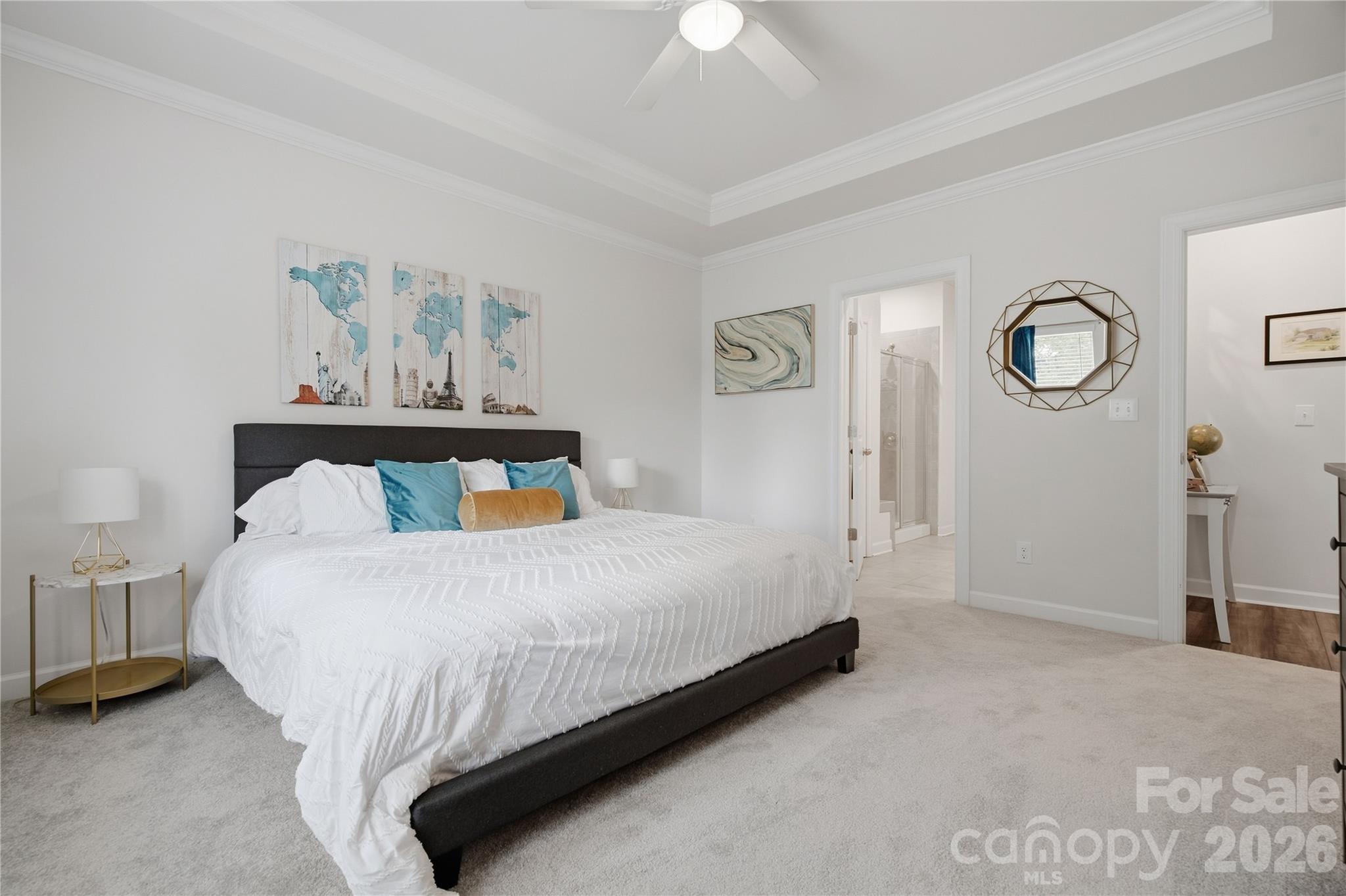 Staged bedroom with a white bed, teal accent pillows, dark headboard, and doorway to an adjoining bathroom.
