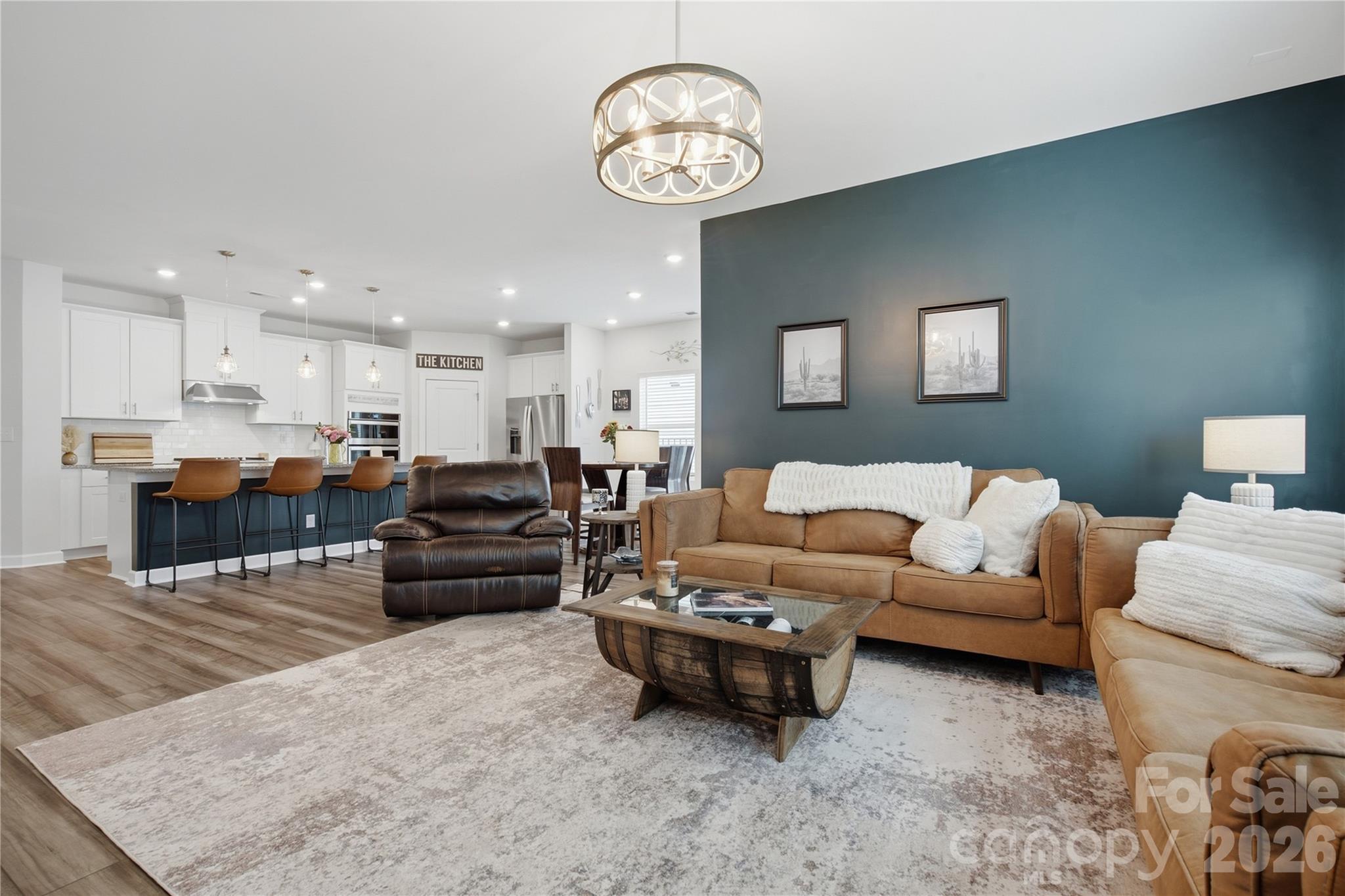 Open-plan living room and kitchen with a tan sectional, teal accent wall, and white cabinetry.