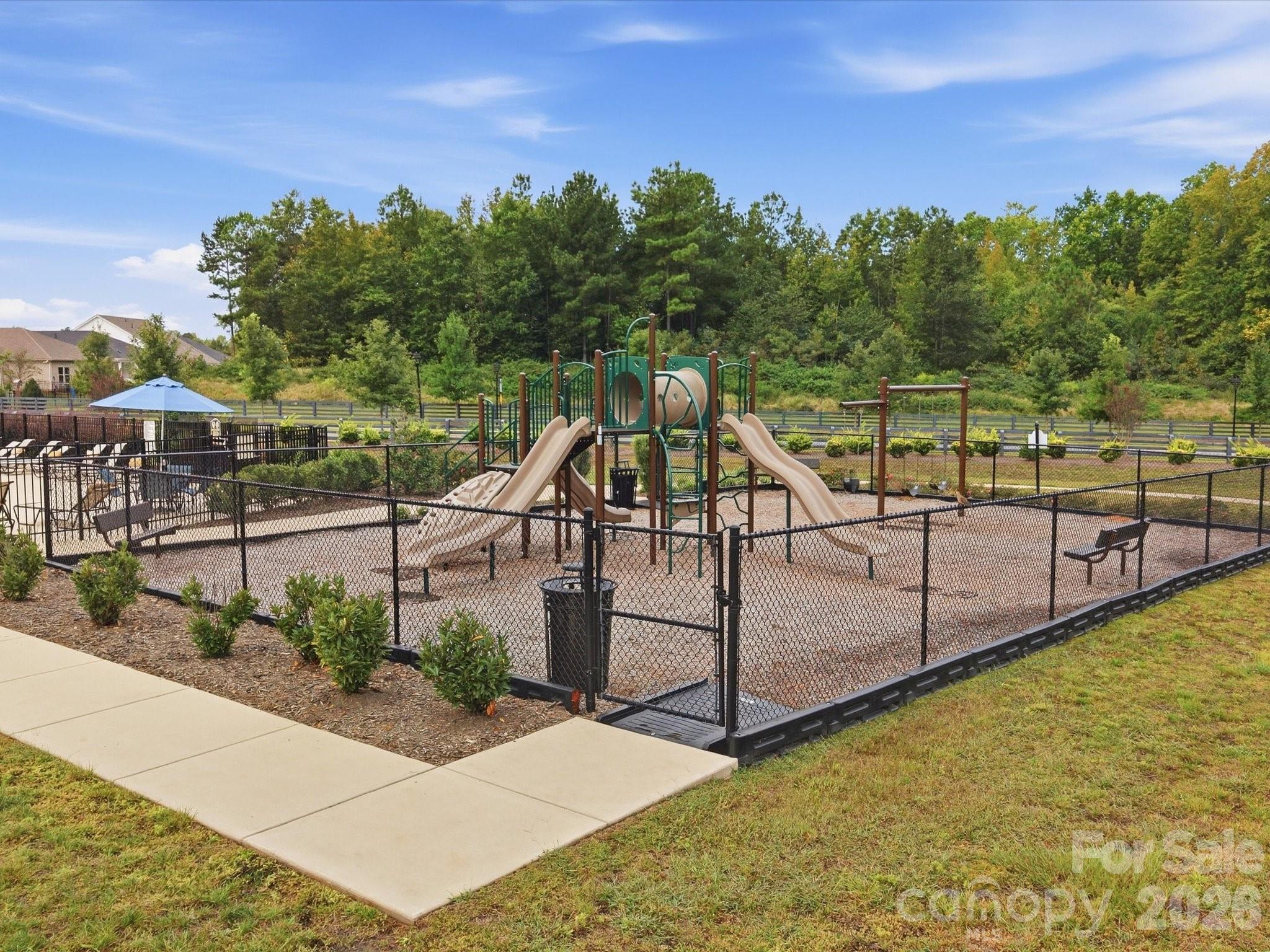 Fenced community playground with a large slide structure, benches, and a shaded seating area.