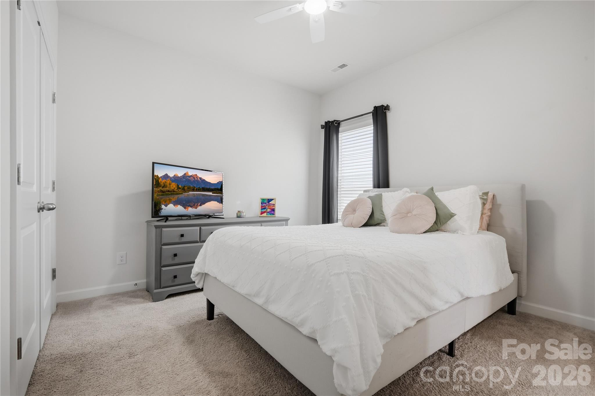 Staged bedroom with a white bed, blush accent pillows, gray dresser, and television.