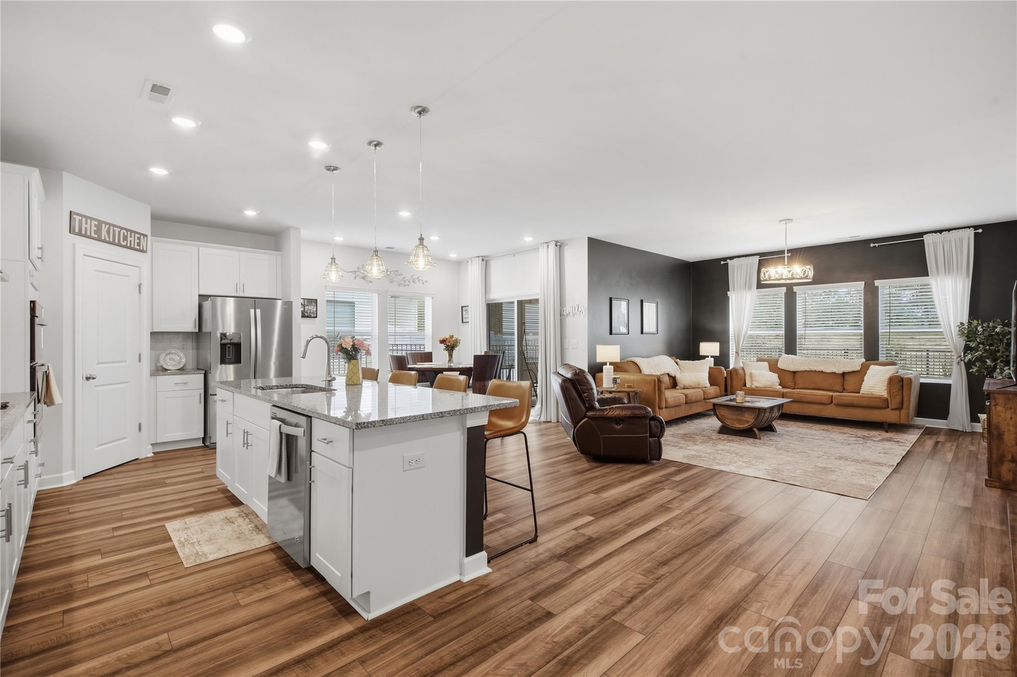 Open kitchen and living room with a granite island, white cabinetry, and tan sofas against a dark accent wall.