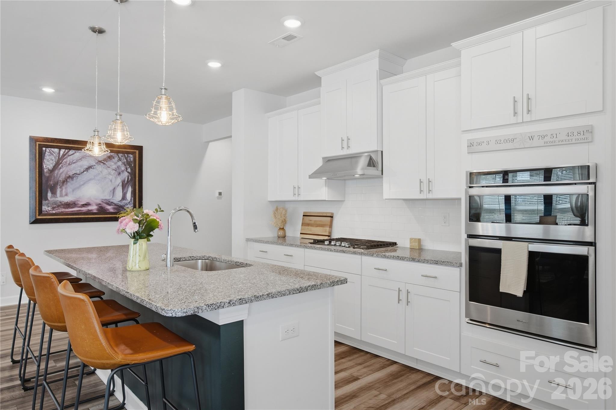 Bright white kitchen with a large island, brown bar stools, pendant lights, and a stainless-steel double oven.
