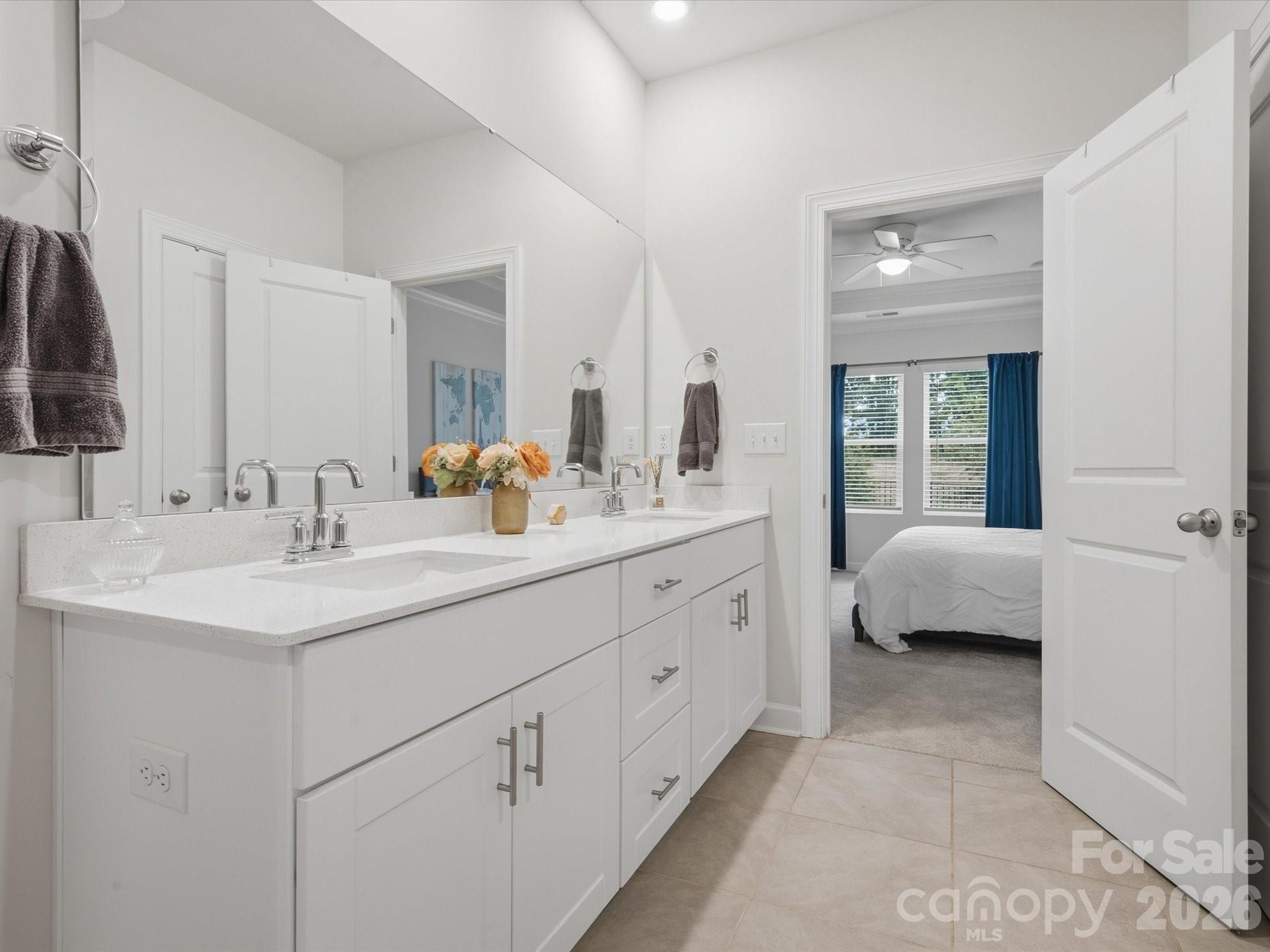 Bathroom with a long white double vanity and mirror, opening onto a bedroom with blue curtains.