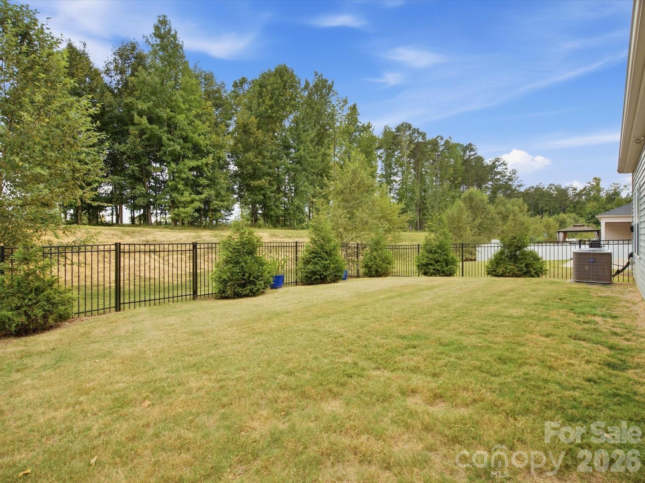 Grassy backyard enclosed by a black metal fence, with shrubs and mature trees.