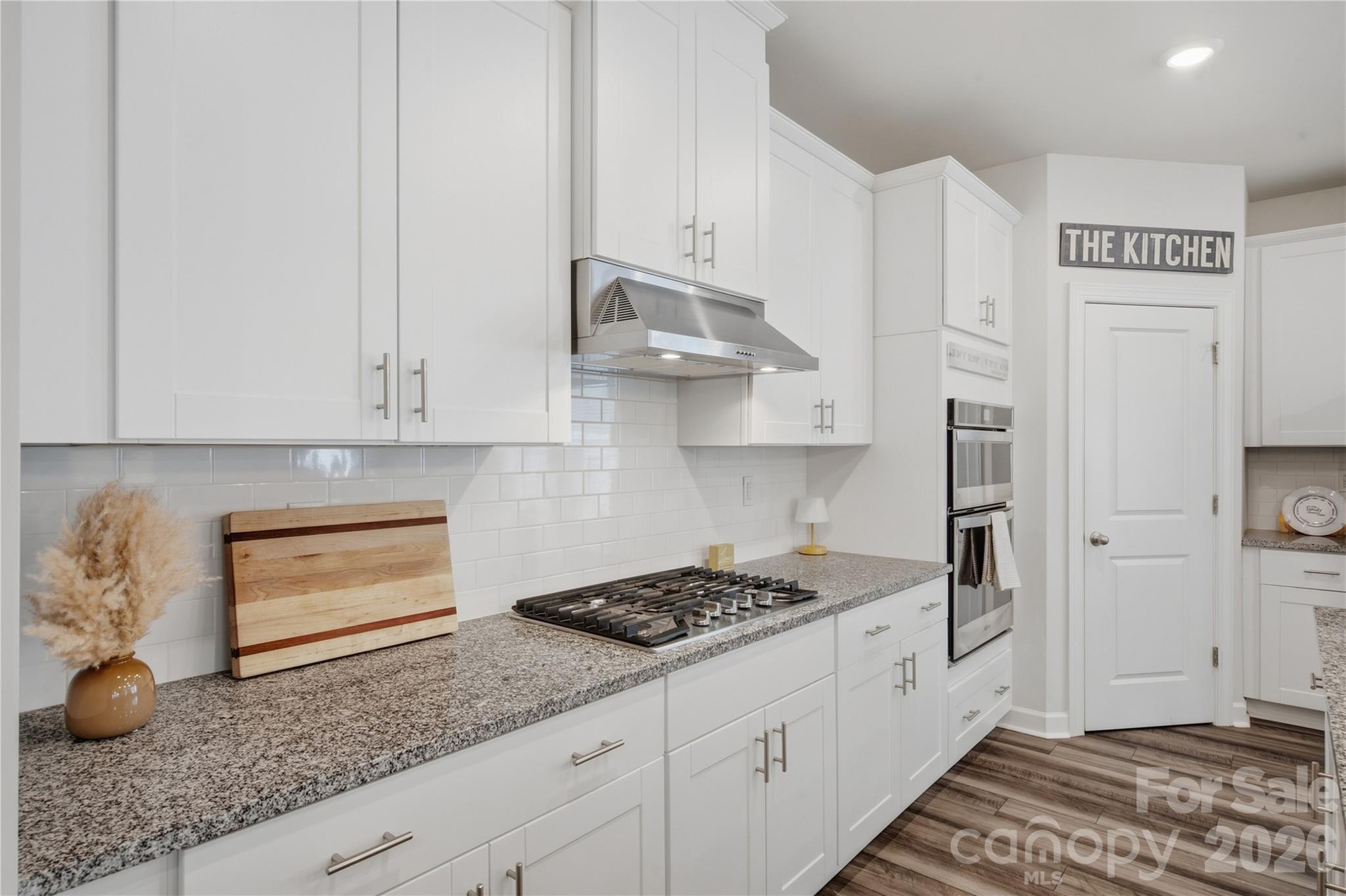 Bright white kitchen with granite counters, gas cooktop, stainless hood, and built-in double oven.