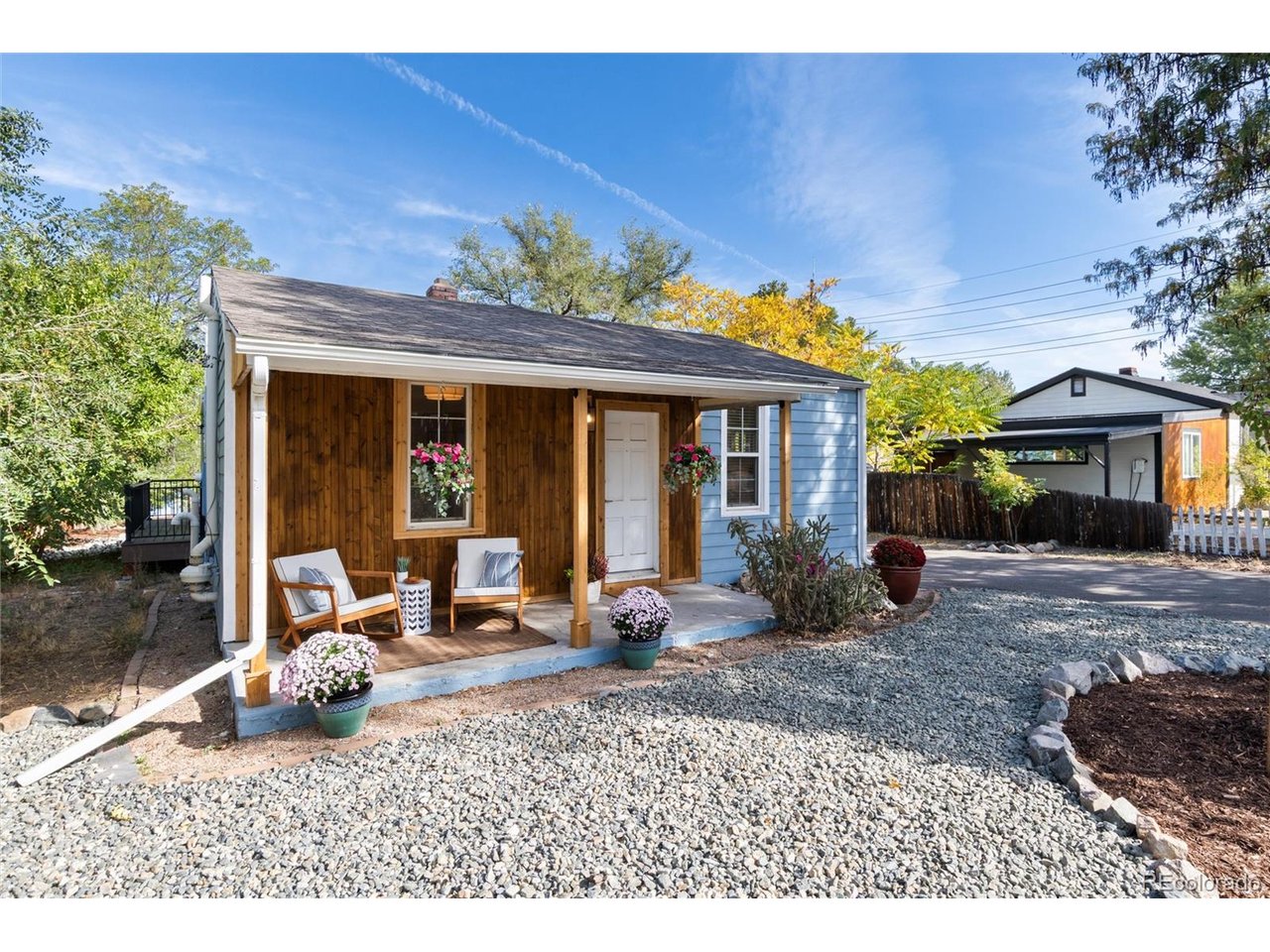 Small house with a wood-paneled porch, blue siding, flower baskets, and a gravel yard.
