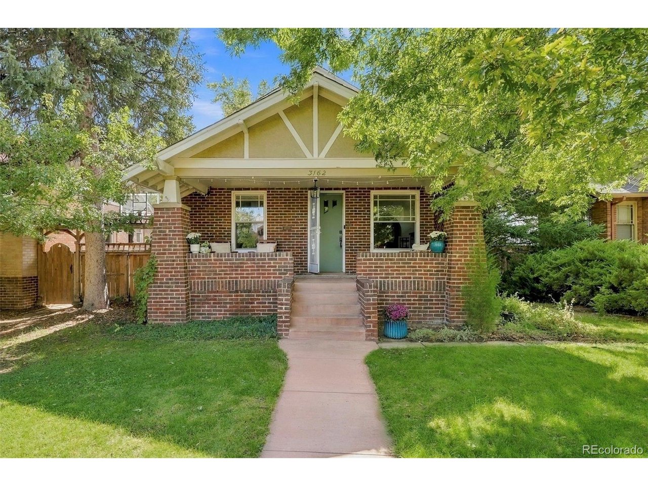 Brick bungalow with a covered porch, green front door, and leafy trees framing the lawn.