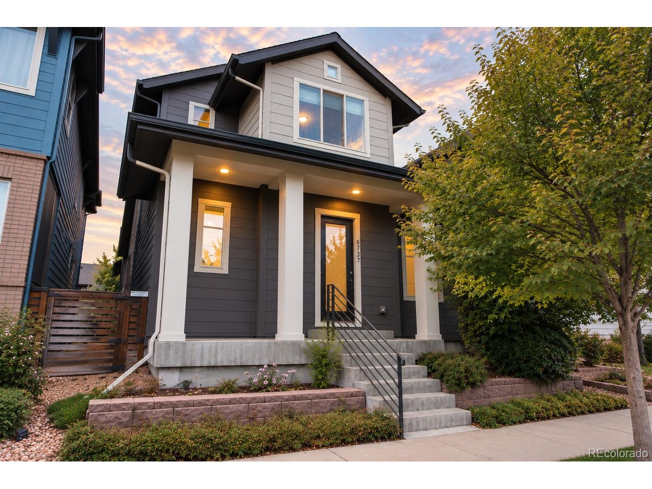 Two-story gray home with a covered porch, concrete steps, and a leafy tree at dusk.