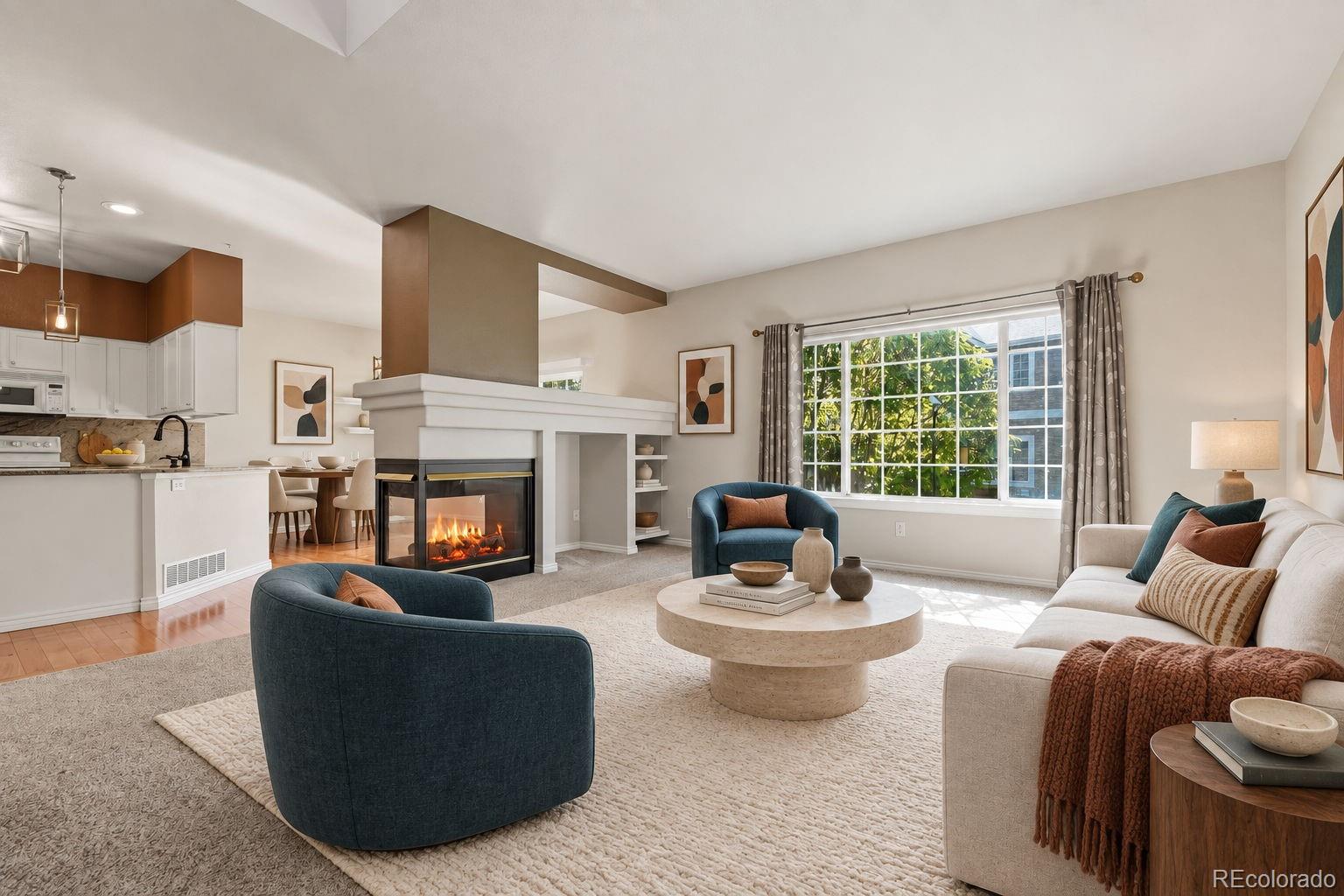 Staged living room with a gas fireplace, blue armchairs, cream sofa, and adjoining kitchen.