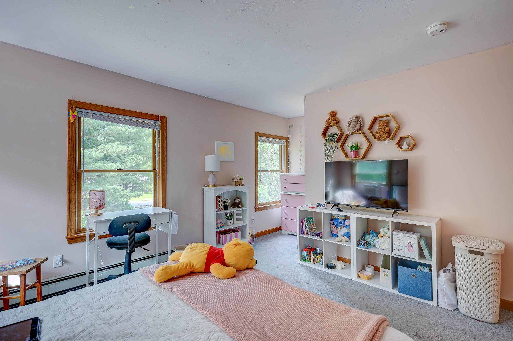 Child's bedroom with a large bed, toy-filled shelving, and a desk beside wide windows.
