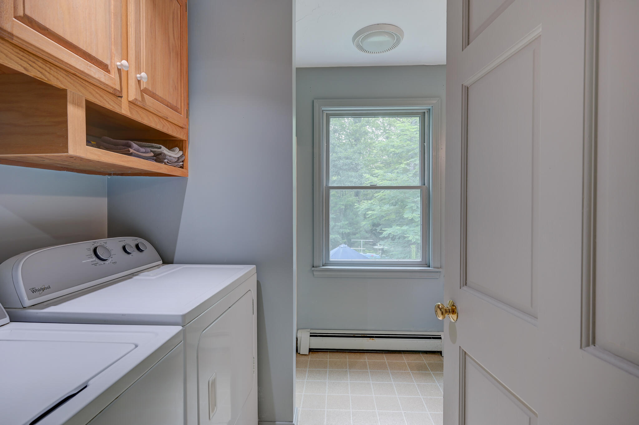 Laundry room with washer and dryer, wooden cabinets, and a window overlooking trees.