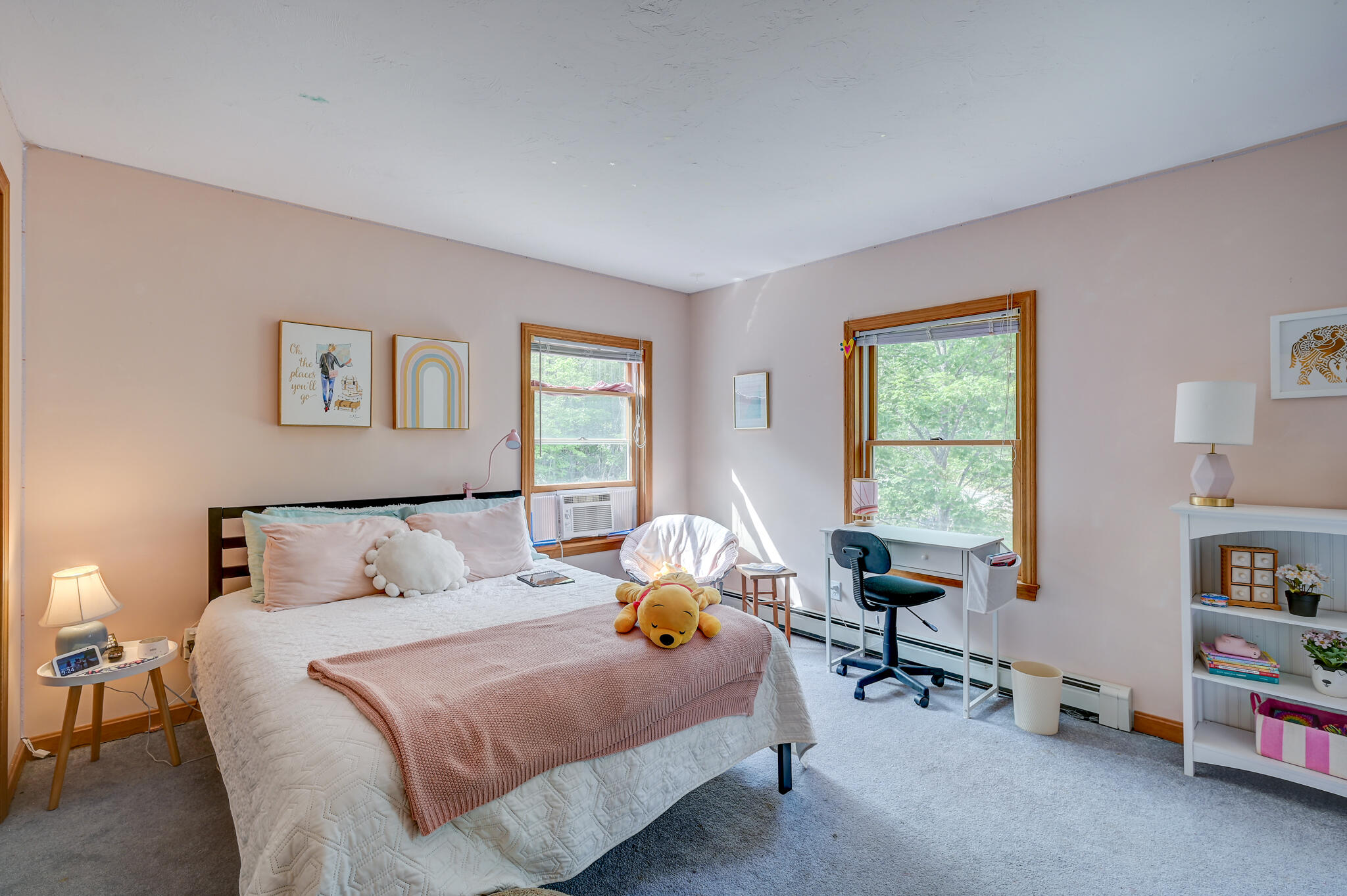 Bedroom with a neatly made bed, two wood-trimmed windows, and a small desk overlooking trees.