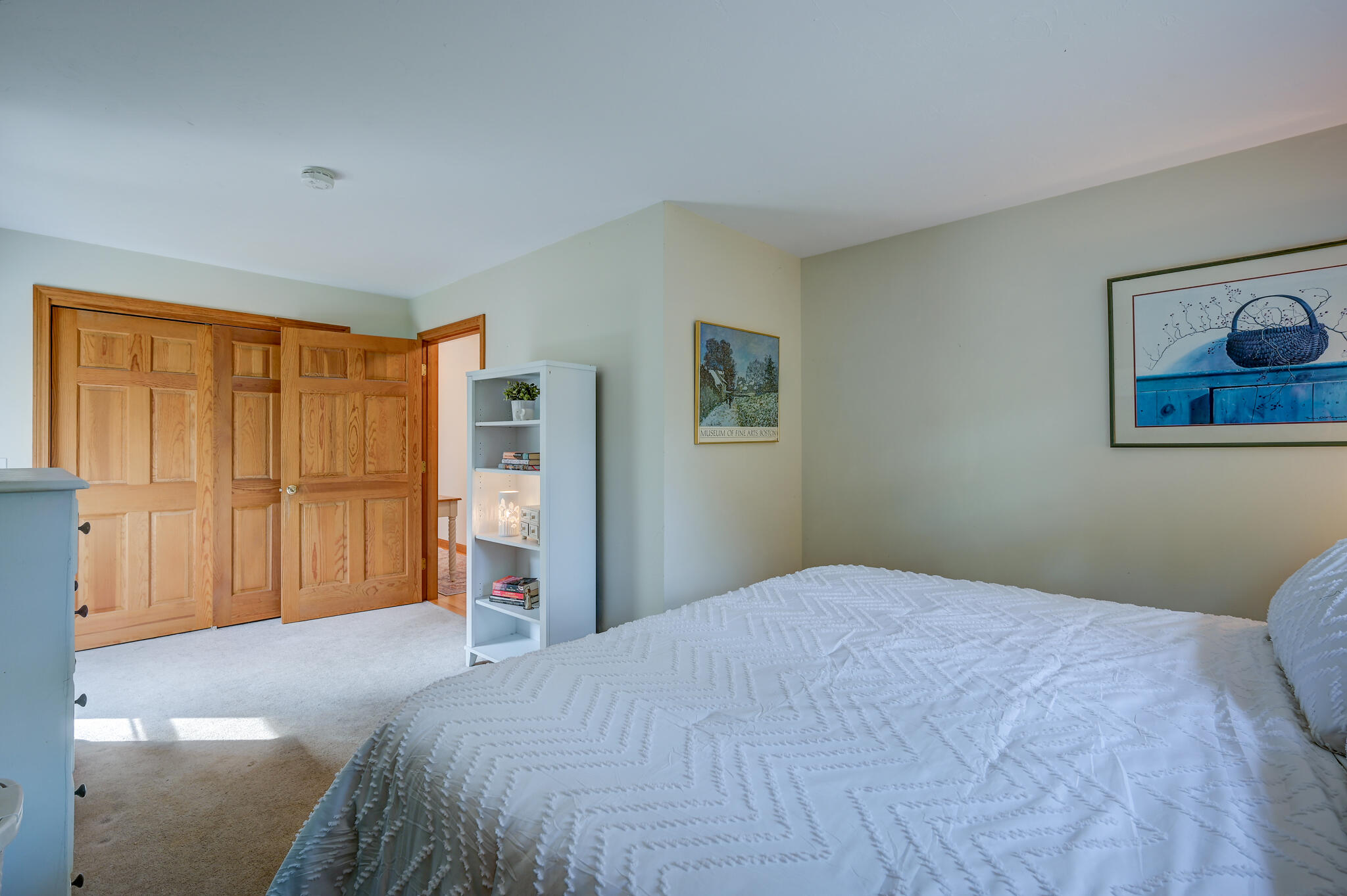 Bedroom with a white quilted bedspread, wooden closet doors, and a narrow bookcase.