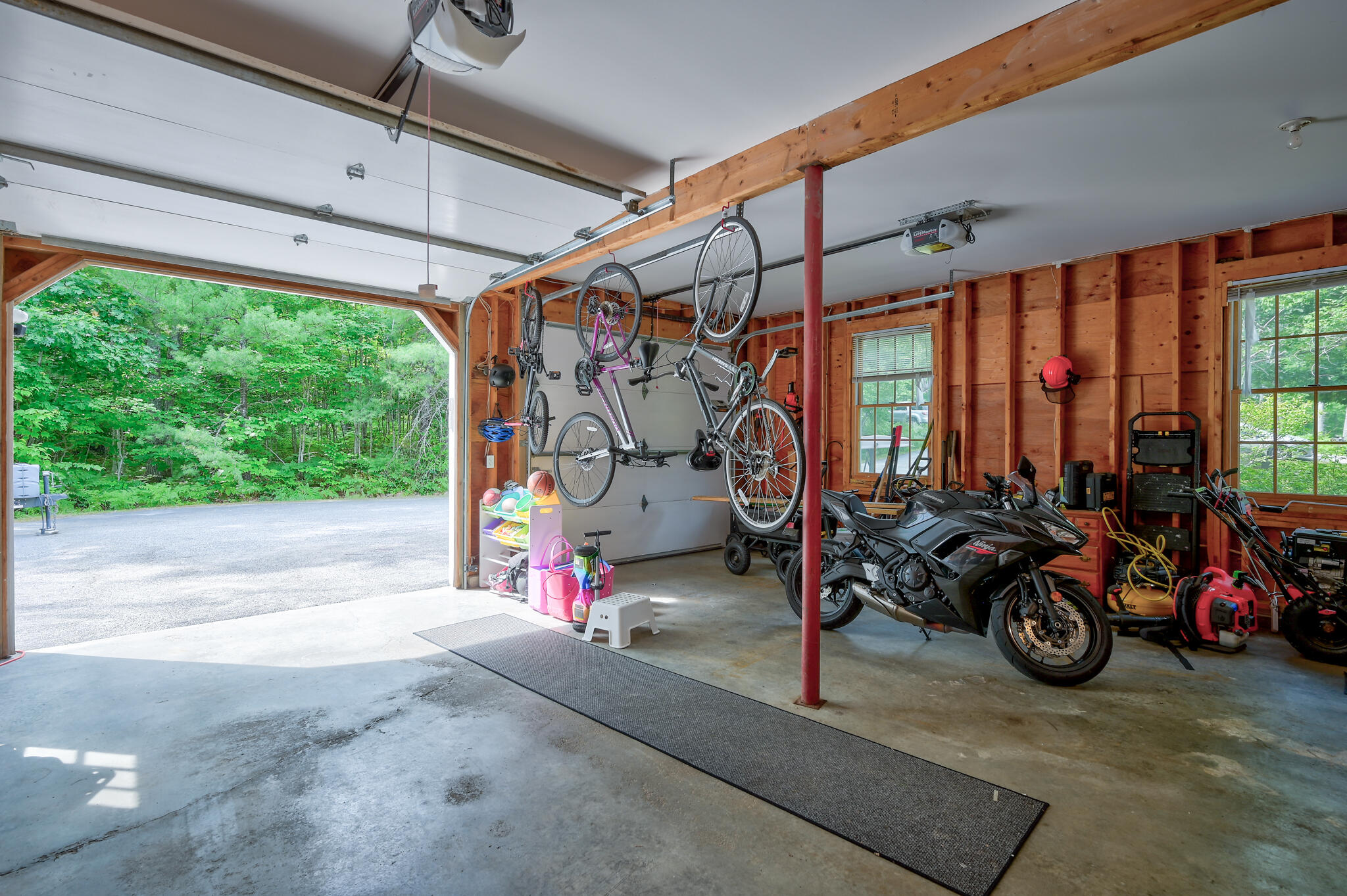 Garage with hanging bicycles, motorcycles, and an open door facing wooded greenery.