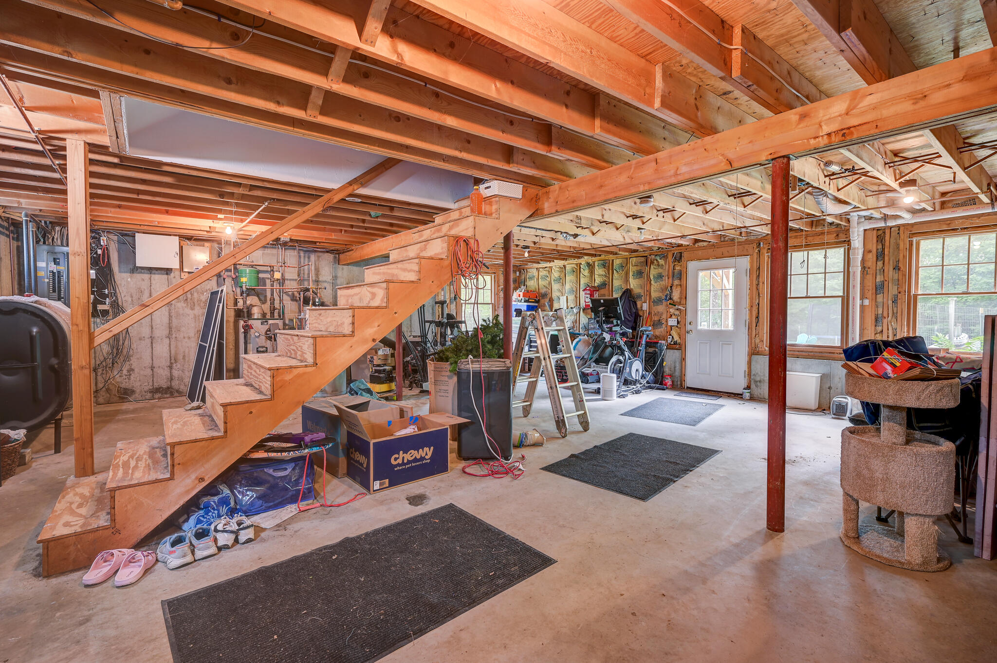 Unfinished basement with exposed beams, a wooden staircase, utility equipment, and storage clutter.