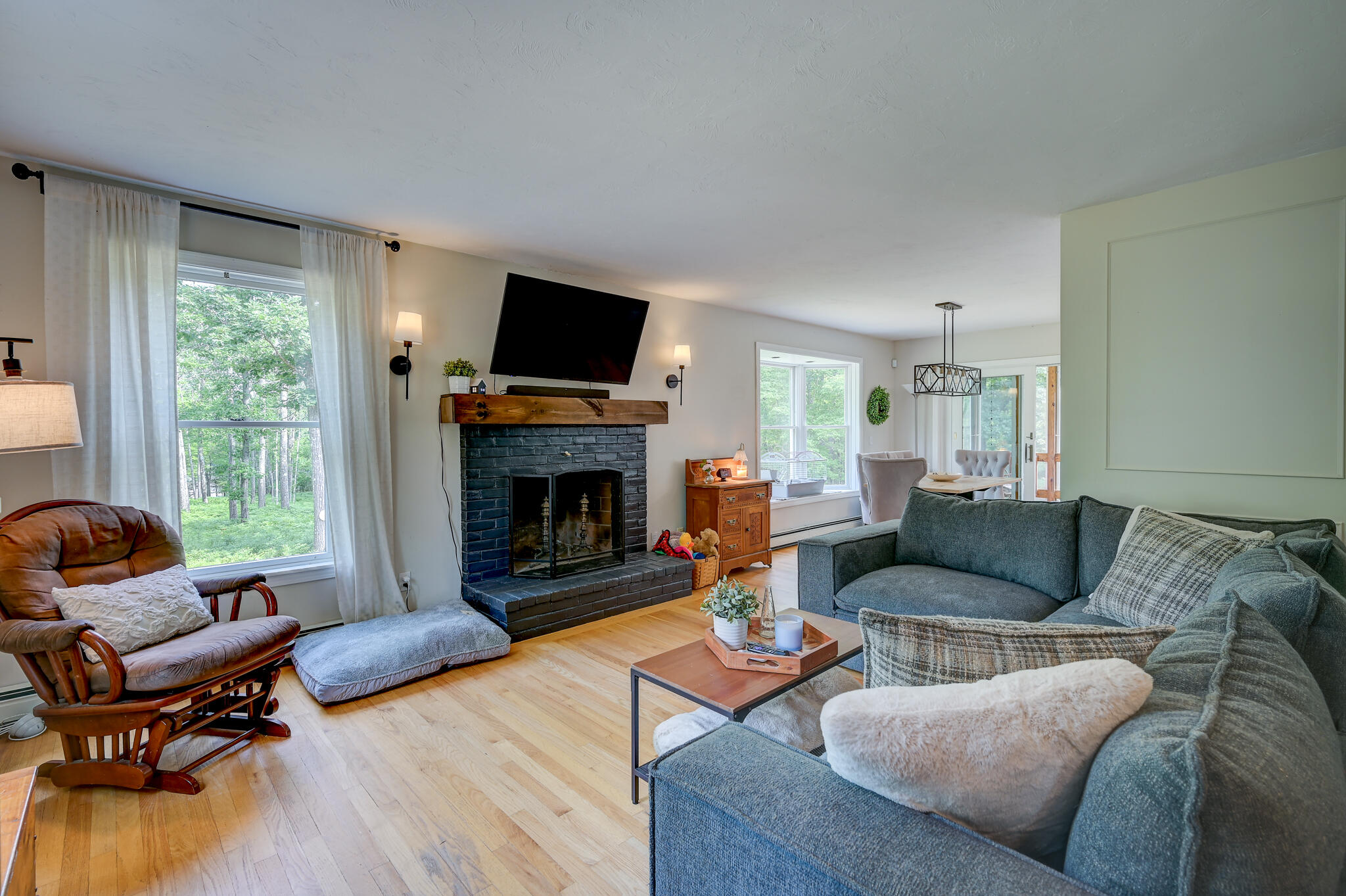 Furnished living room with a brick fireplace, large windows overlooking trees, and a blue sectional sofa.