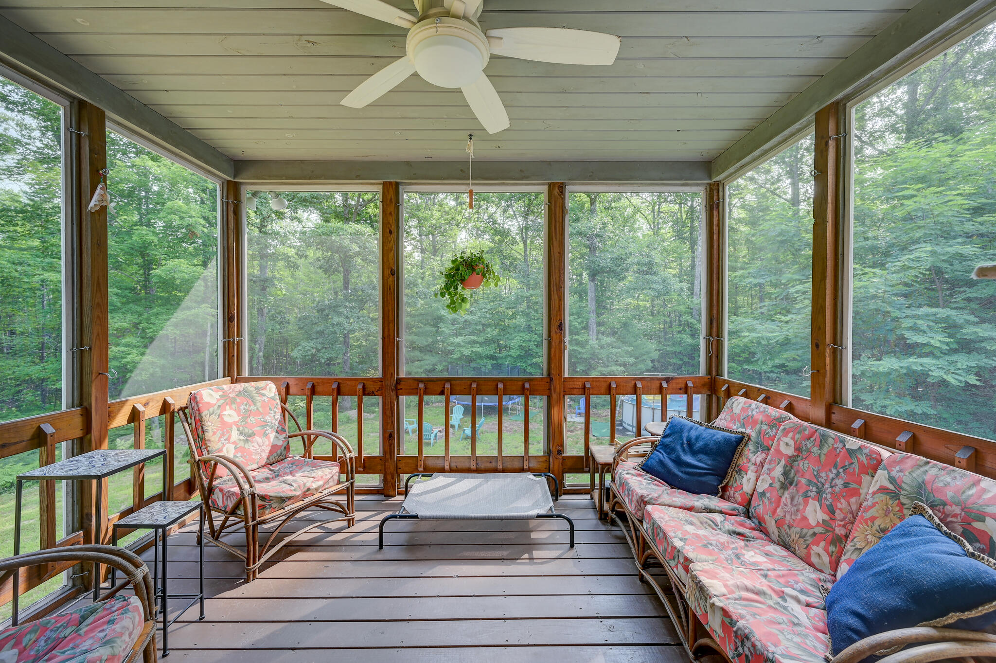 Screened porch with floral seating, ceiling fan, and views of surrounding trees.