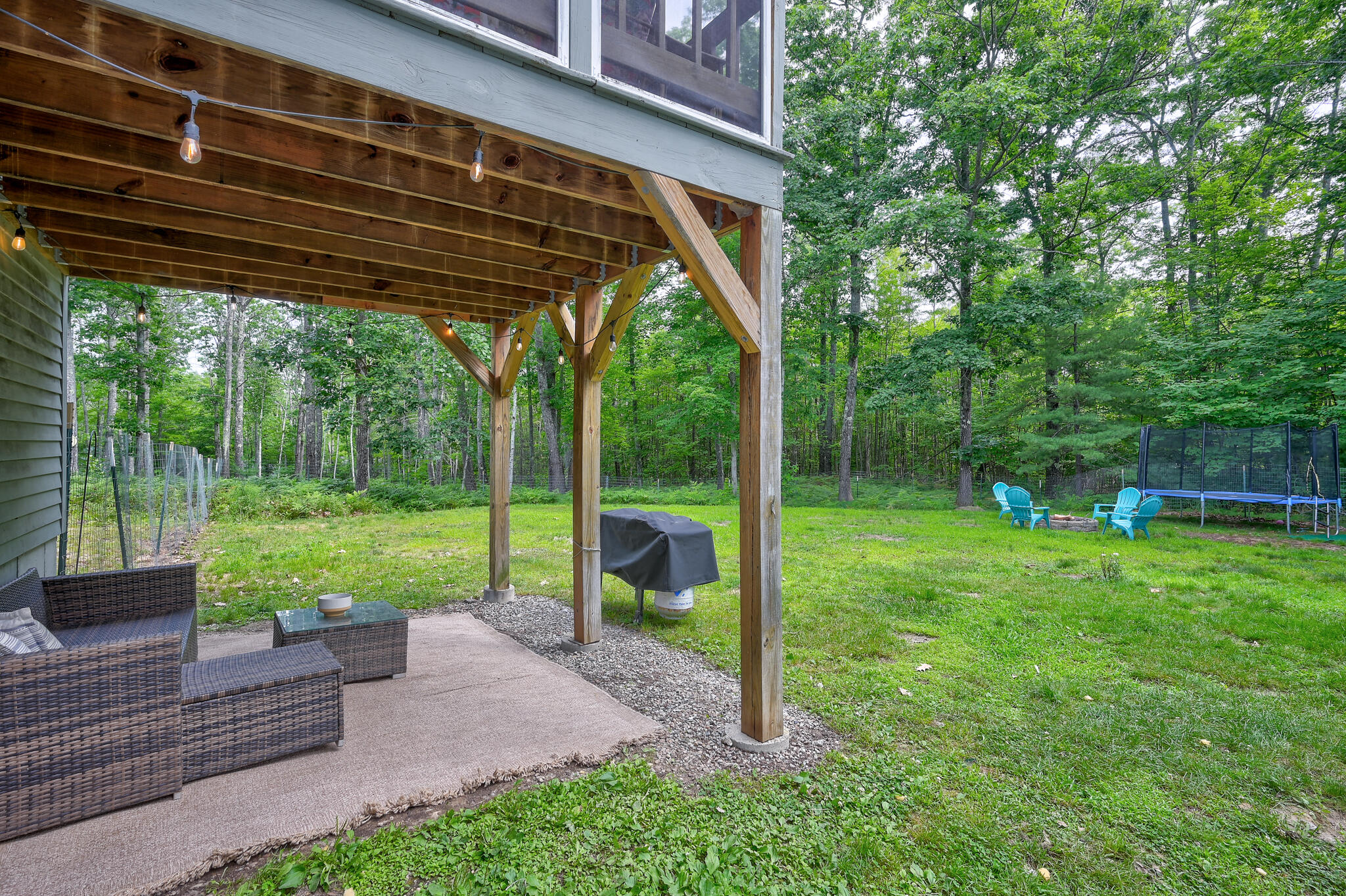 Covered wooden patio overlooking a grassy, wooded yard with outdoor seating and a trampoline.