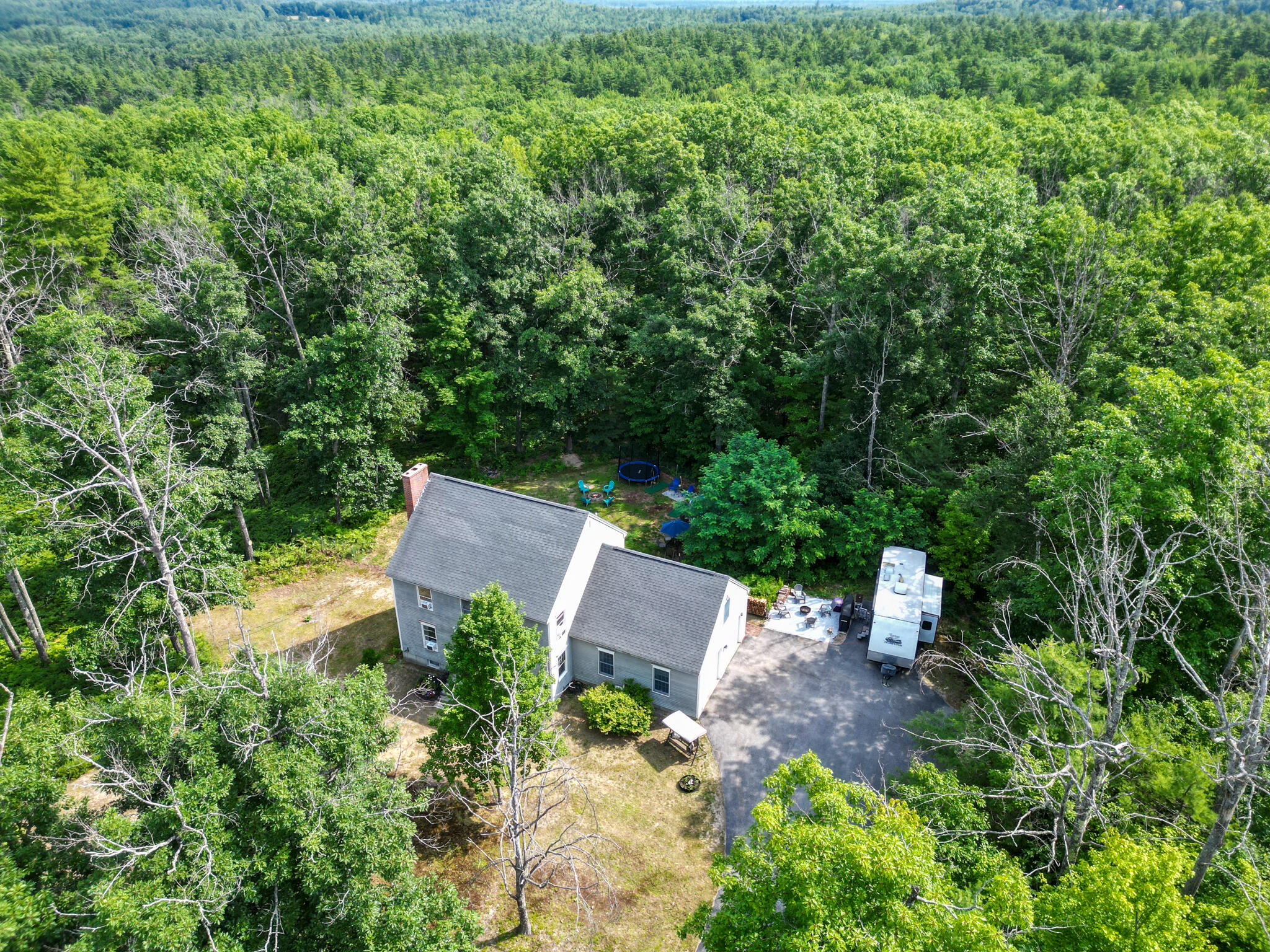 Aerial view of a gray home surrounded by woods, with a driveway and backyard trampoline.