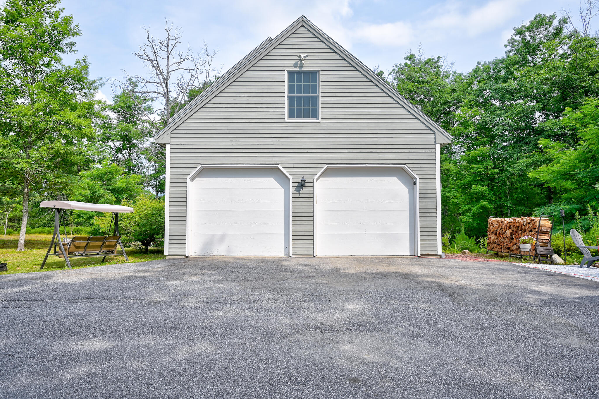 Gray gabled garage with two white doors, a paved driveway, and a swing beneath leafy trees.