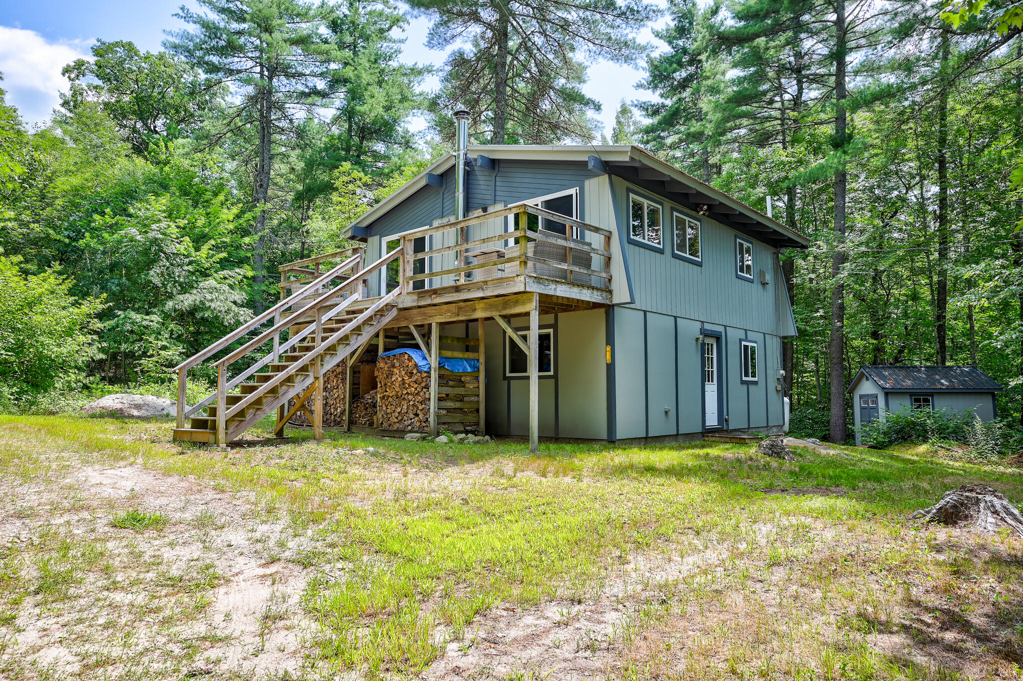 Blue-gray wooded home with a raised wooden deck, exterior stairs, and stacked firewood.