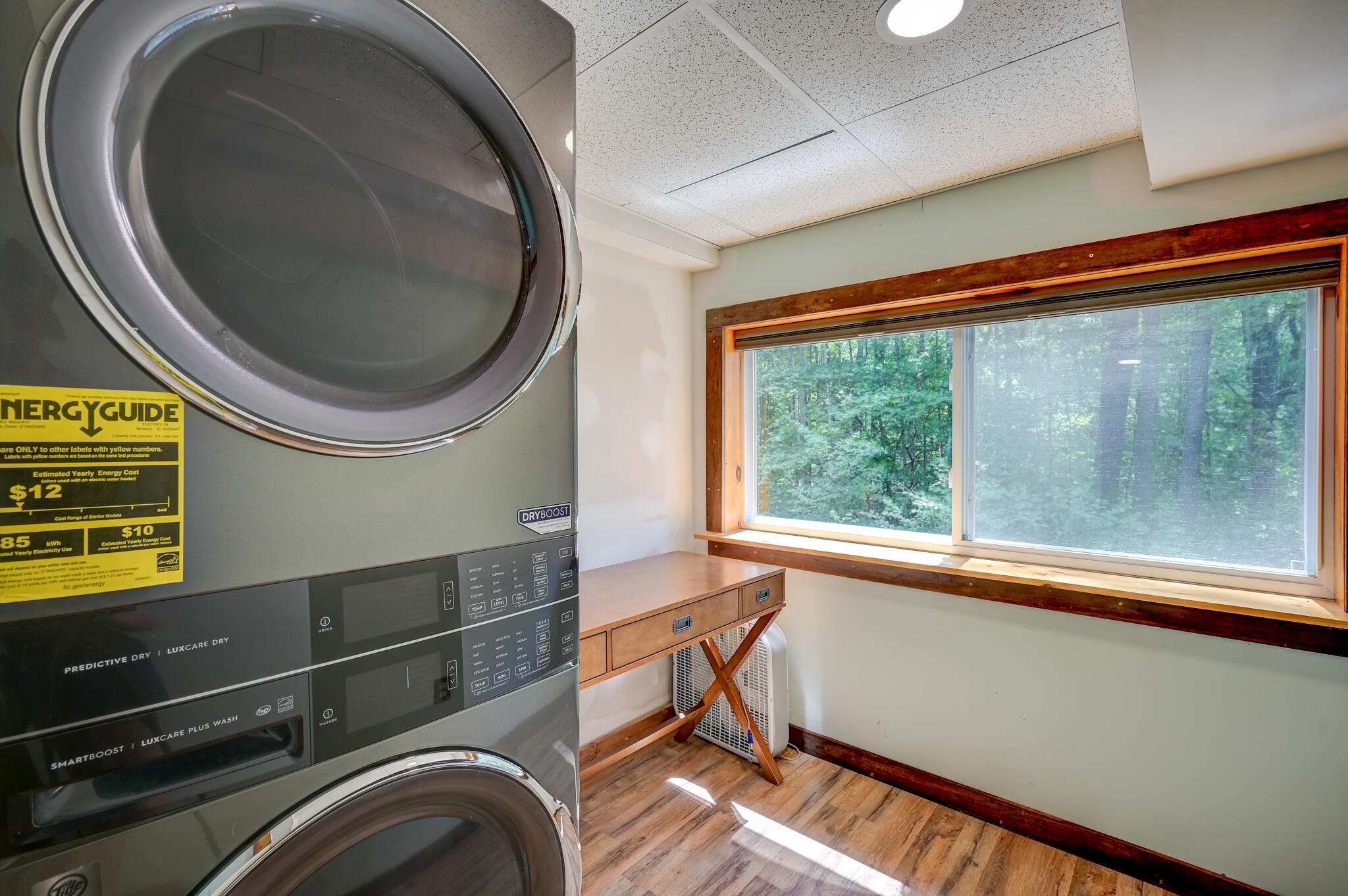 Laundry room with stacked washer and dryer beside a wide window overlooking trees.