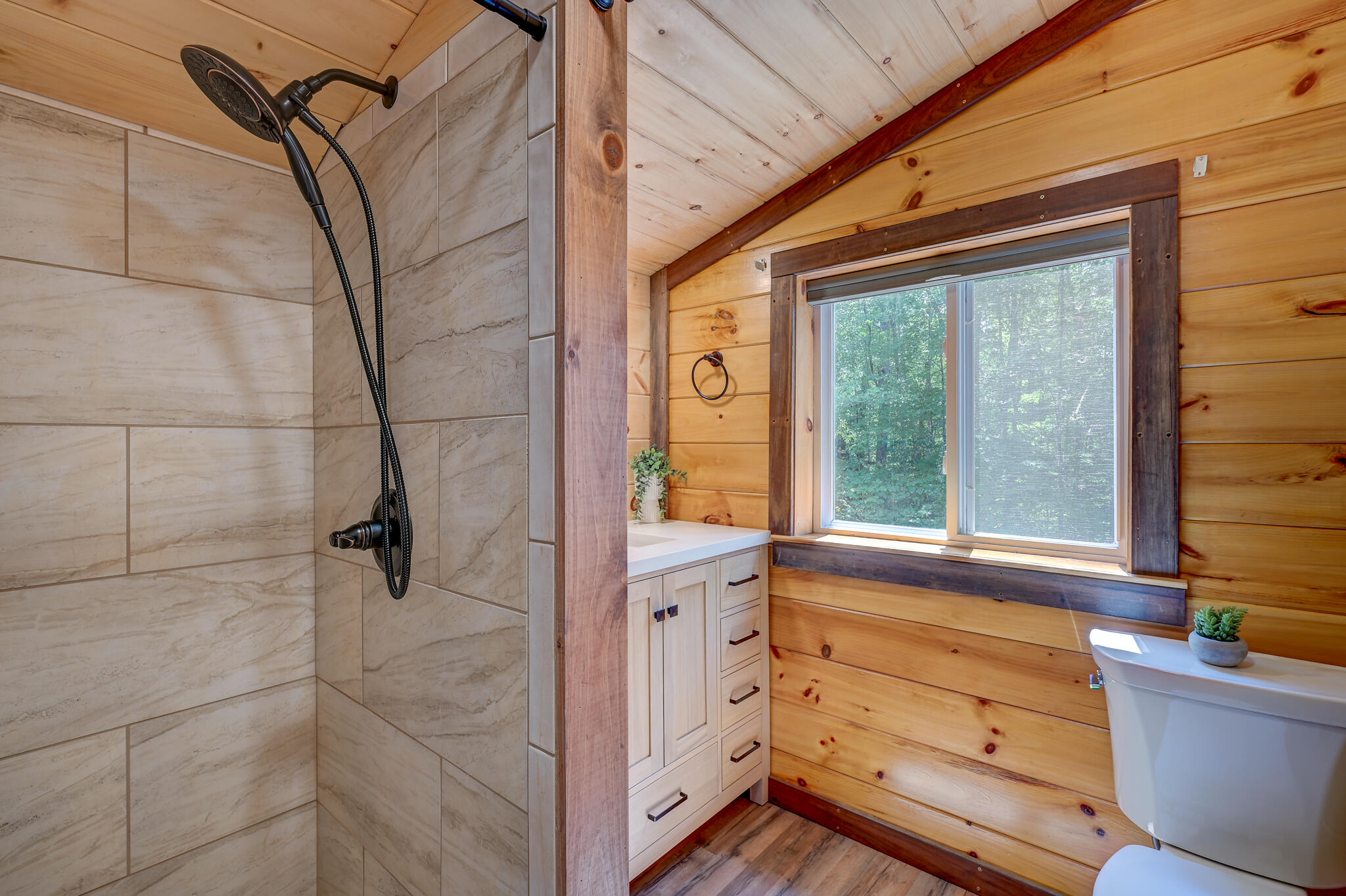 Bathroom with a tiled shower, wood-paneled walls, and a window overlooking trees.