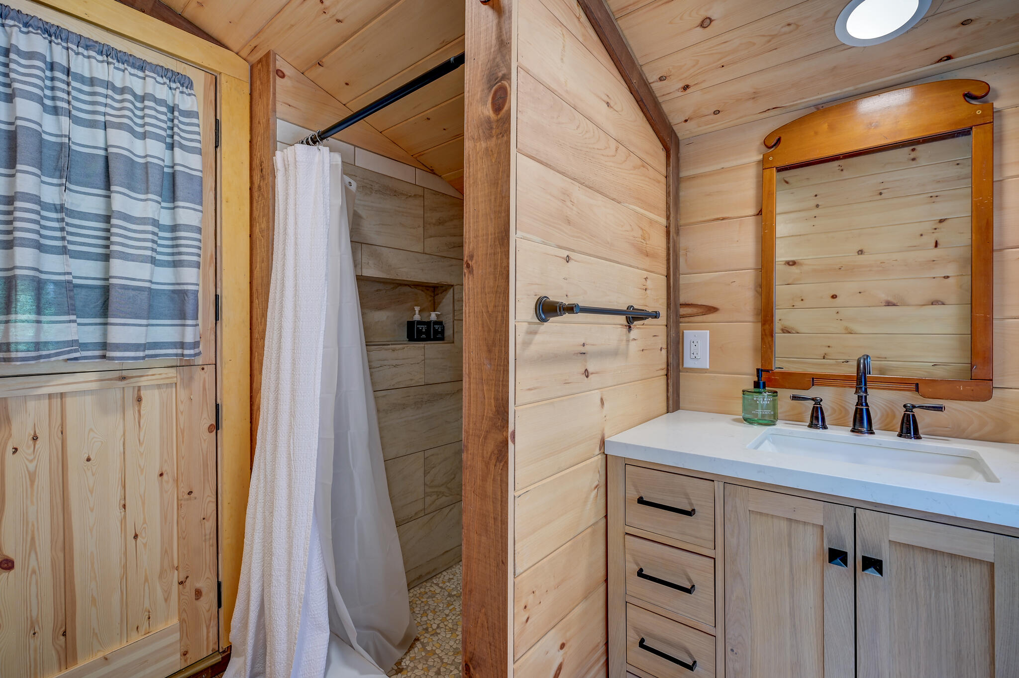 Bathroom with a wood-paneled shower, white curtain, and pale wood vanity beneath a framed mirror.