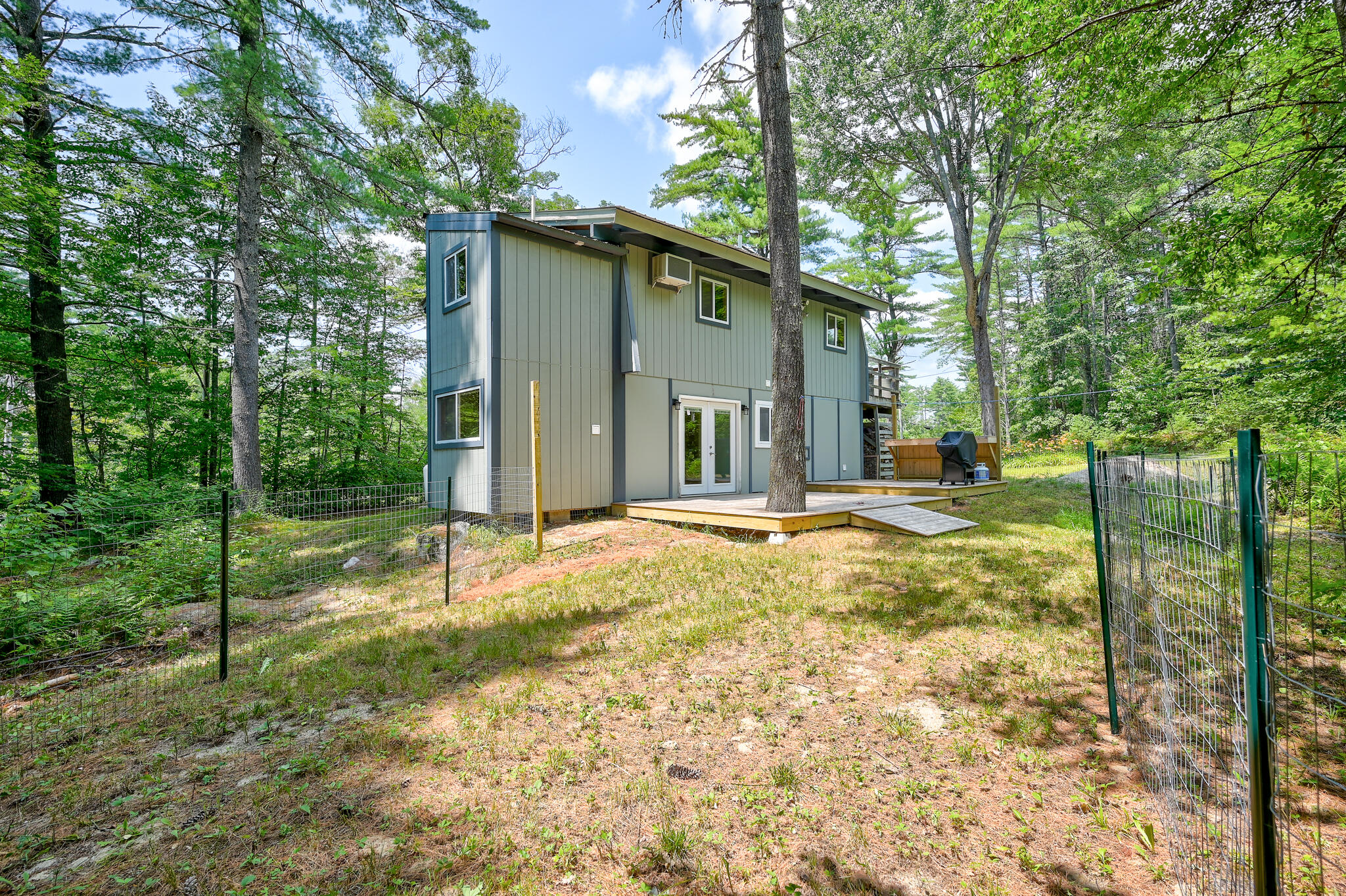 Gray two-story home with a low wooden deck, surrounded by trees and fencing.