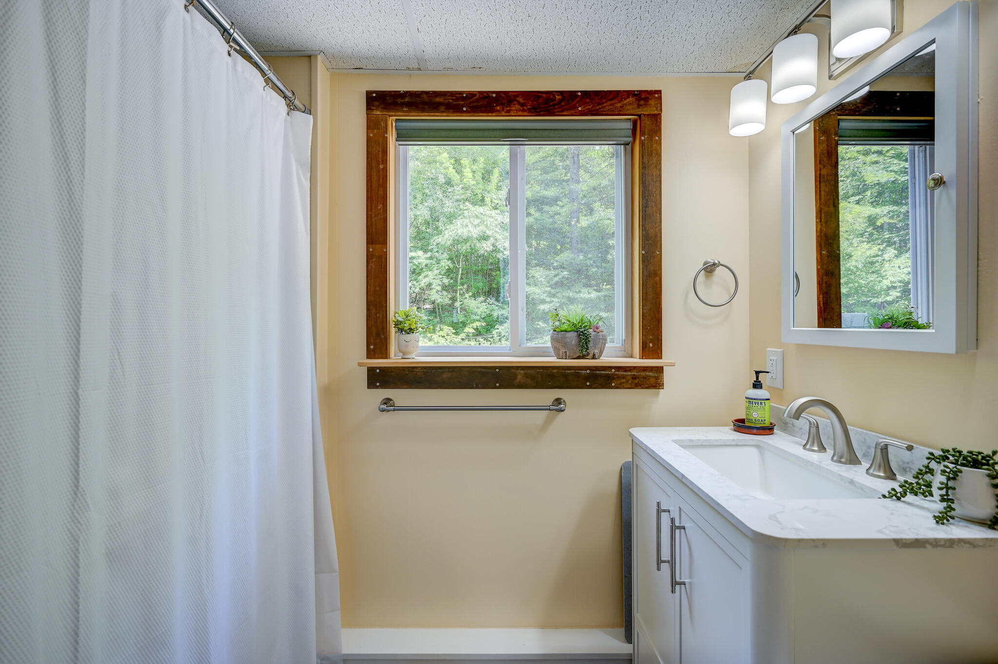 Bathroom with a white vanity, shower curtain, and wood-trimmed window overlooking trees.